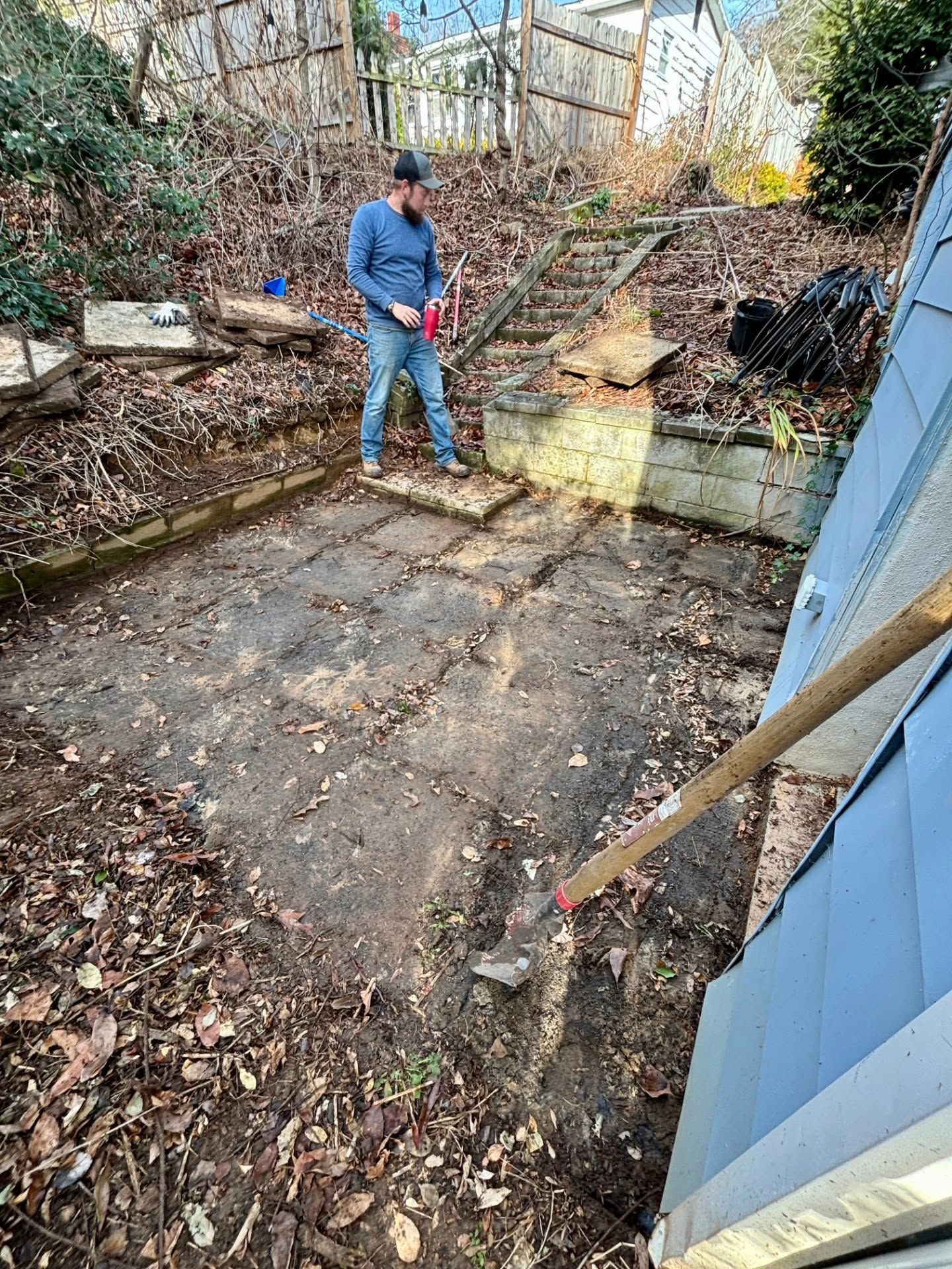 Person in blue jacket working in a leaf-covered backyard beside a stone retaining wall and blue shed