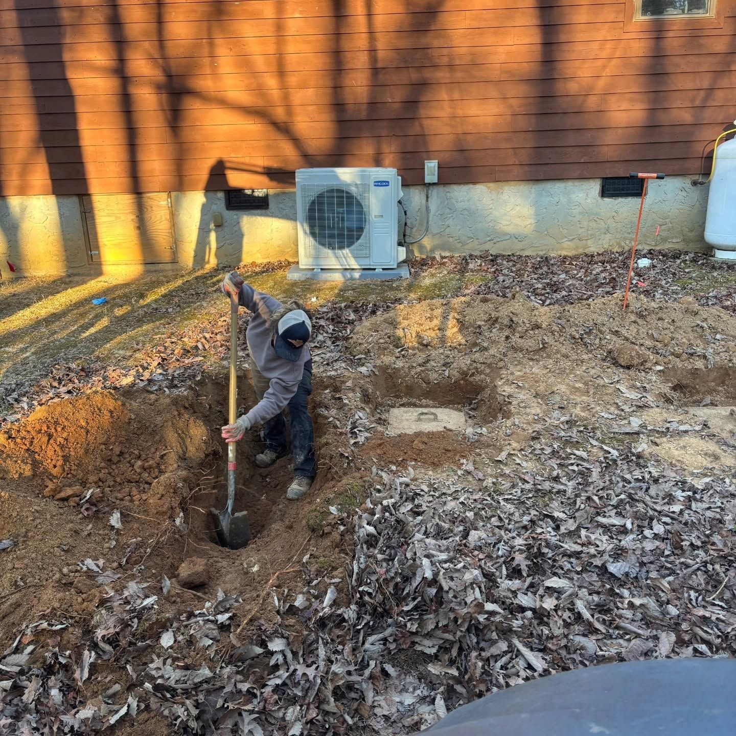 Person digging a hole in a leaf-covered backyard beside a brick house and AC unit