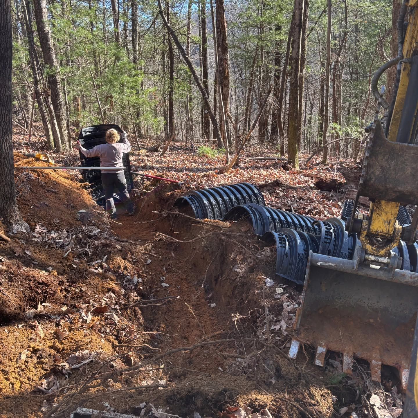 Tractor digging a trench in a wooded area, with a worker and black drainage pipes along the ditch.