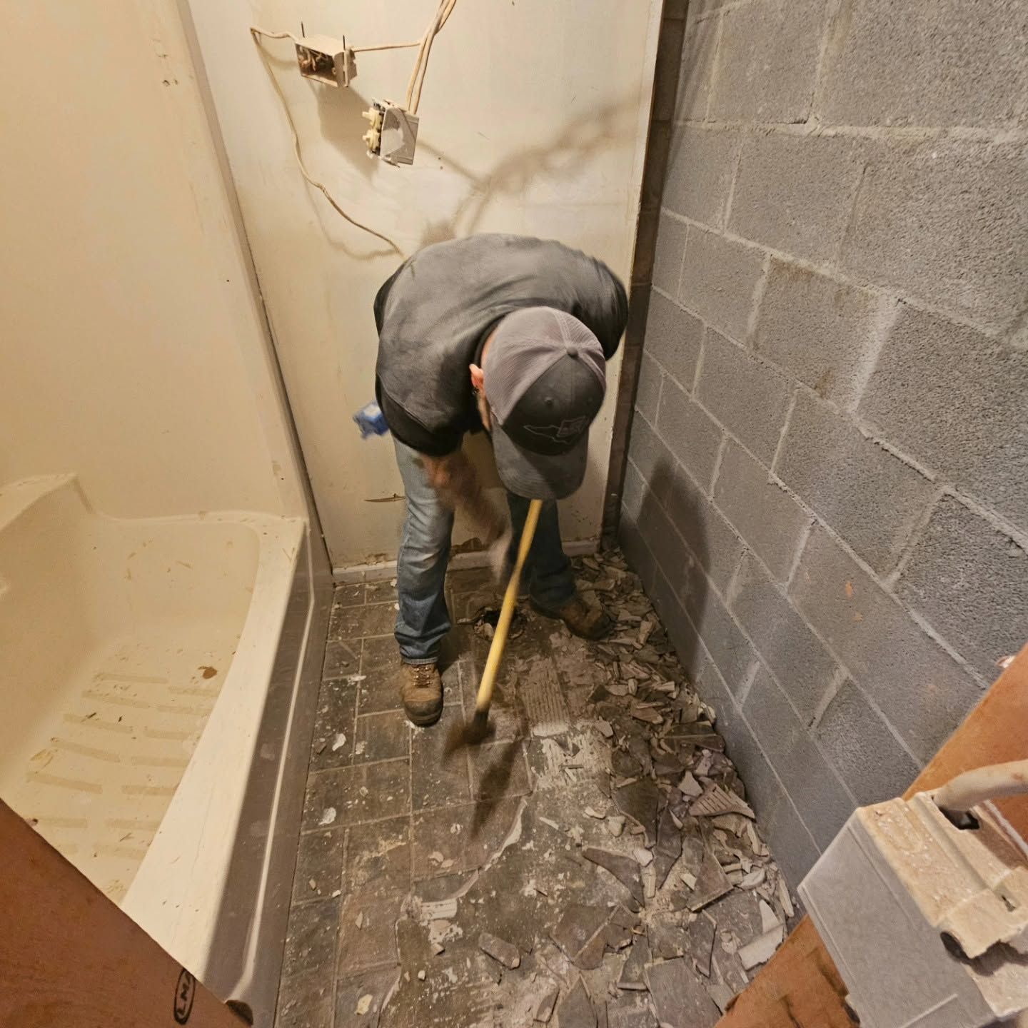 Worker shoveling debris in a narrow bathroom under demolition, with a bathtub on the left and cinder block wall on the right
