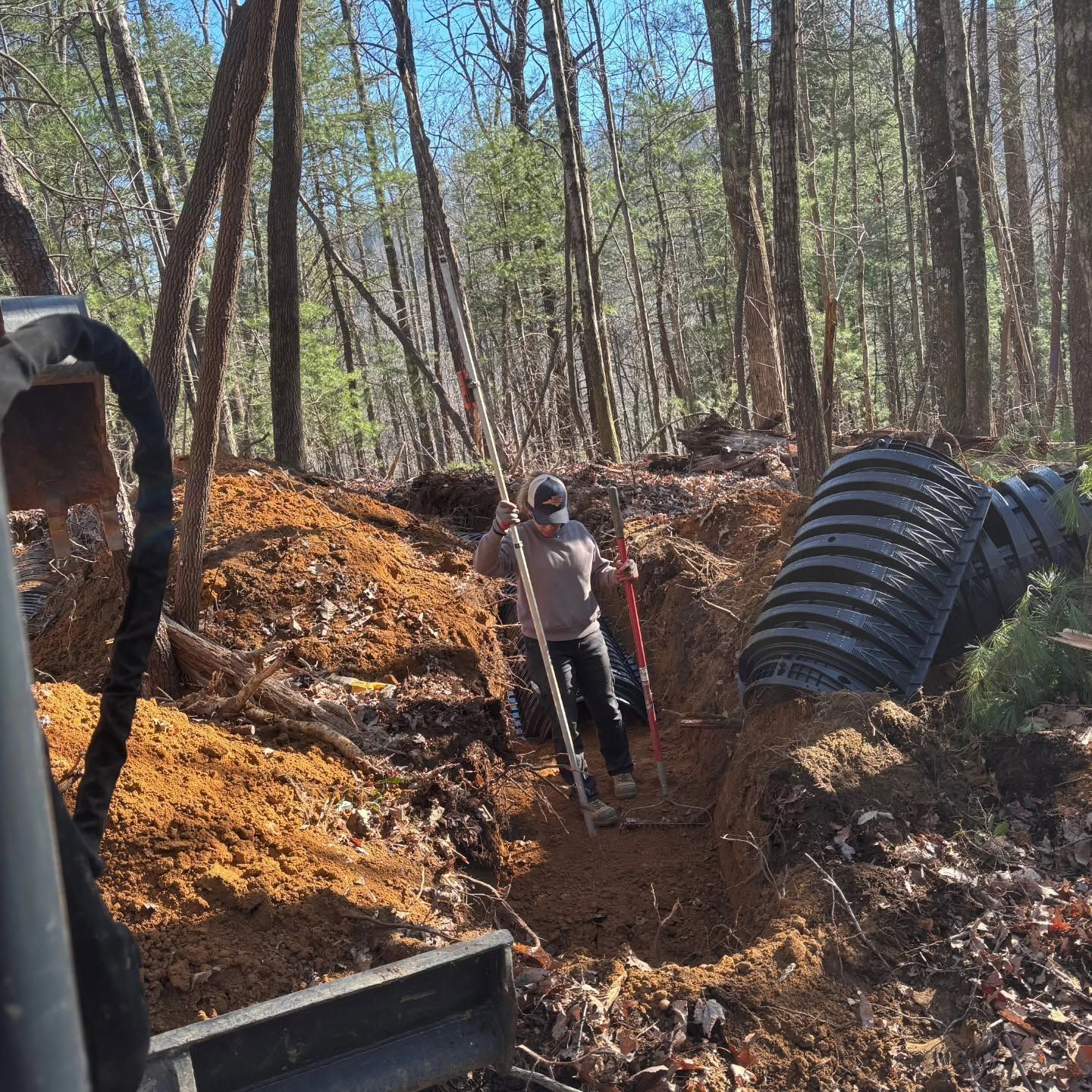 Worker standing in a narrow trench beside large black drainage pipes in a wooded construction area