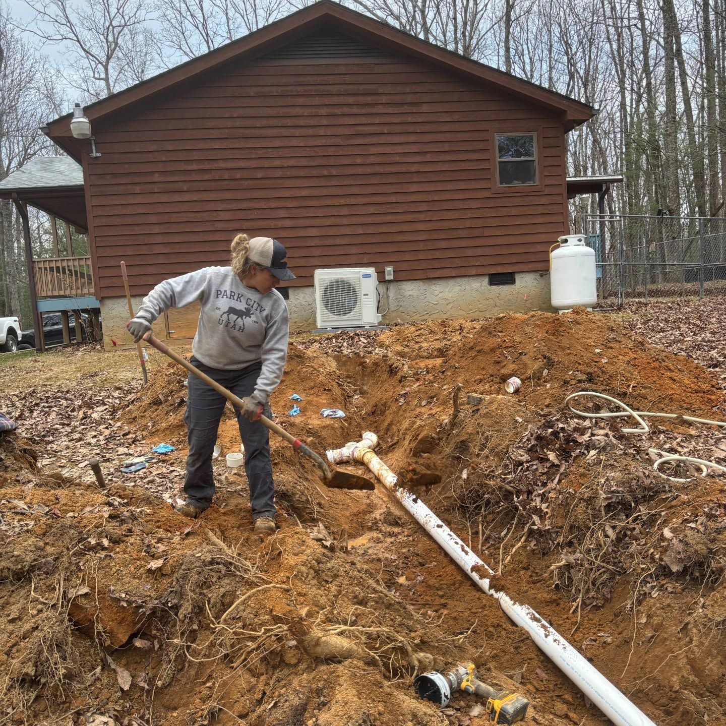 Person working in a muddy excavation beside a brown house with white pipes and a tank.