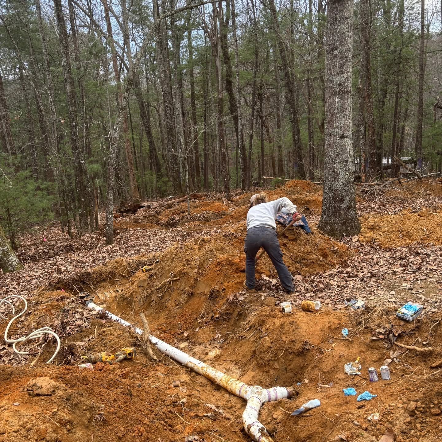 Person digging in a muddy forest excavation site near exposed pipes and blue debris.