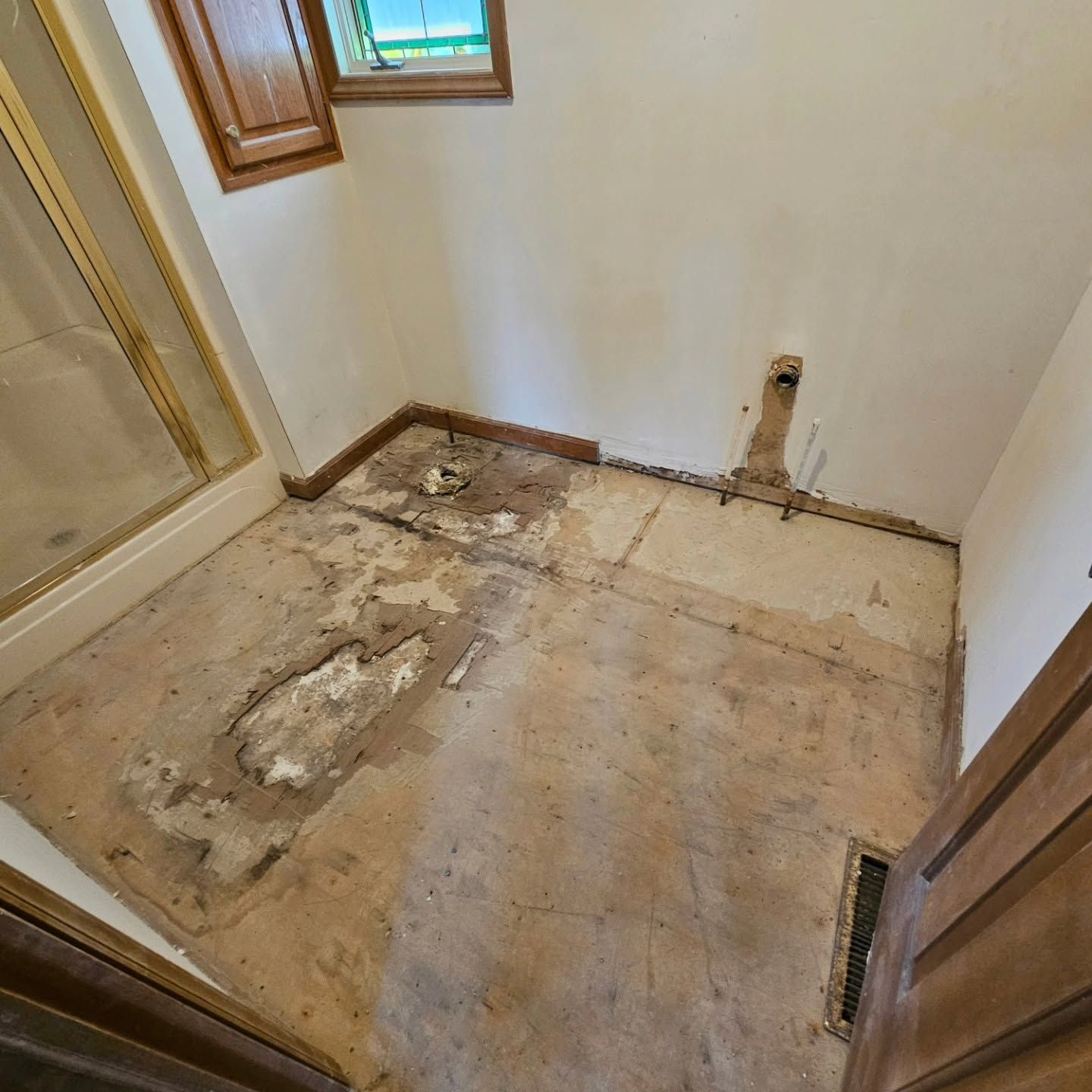 Small unfinished bathroom with damaged subfloor, exposed patching, white walls, and wood trim around windows and door.
