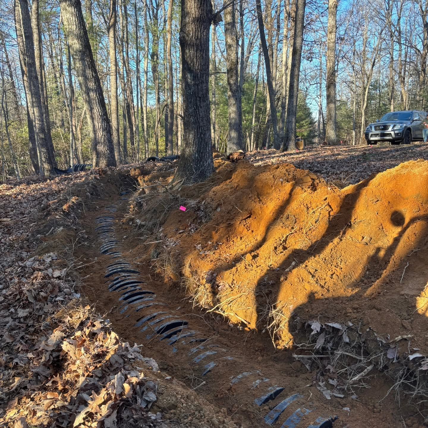 Steep orange dirt embankment with tire tracks and trees in a wooded area, car parked at top right