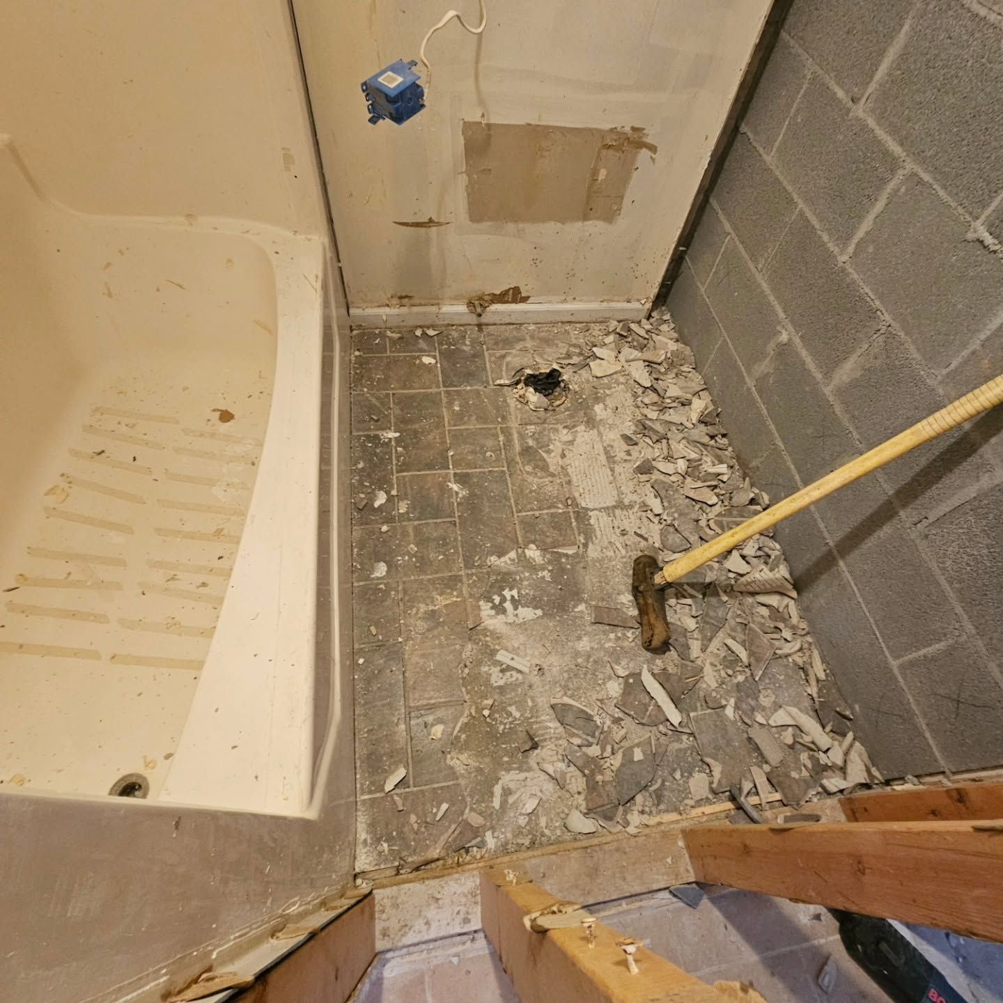 Damaged bathroom floor with broken tiles, debris, a shower tub edge, and a hammer on the floor
