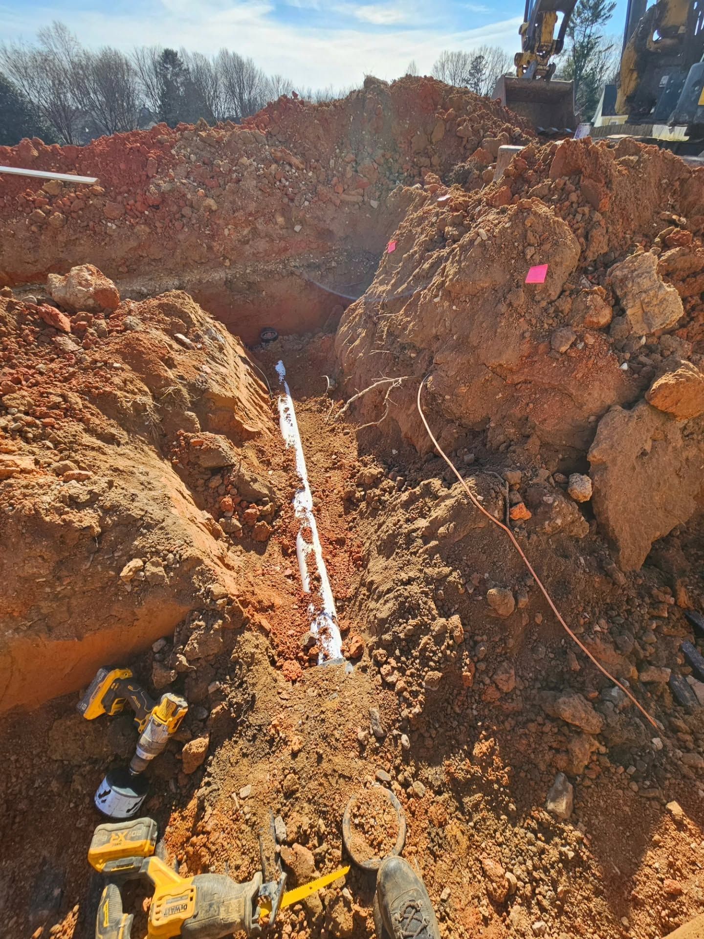 Trenched construction site with exposed dirt, a white pipe, and heavy equipment on a muddy slope.