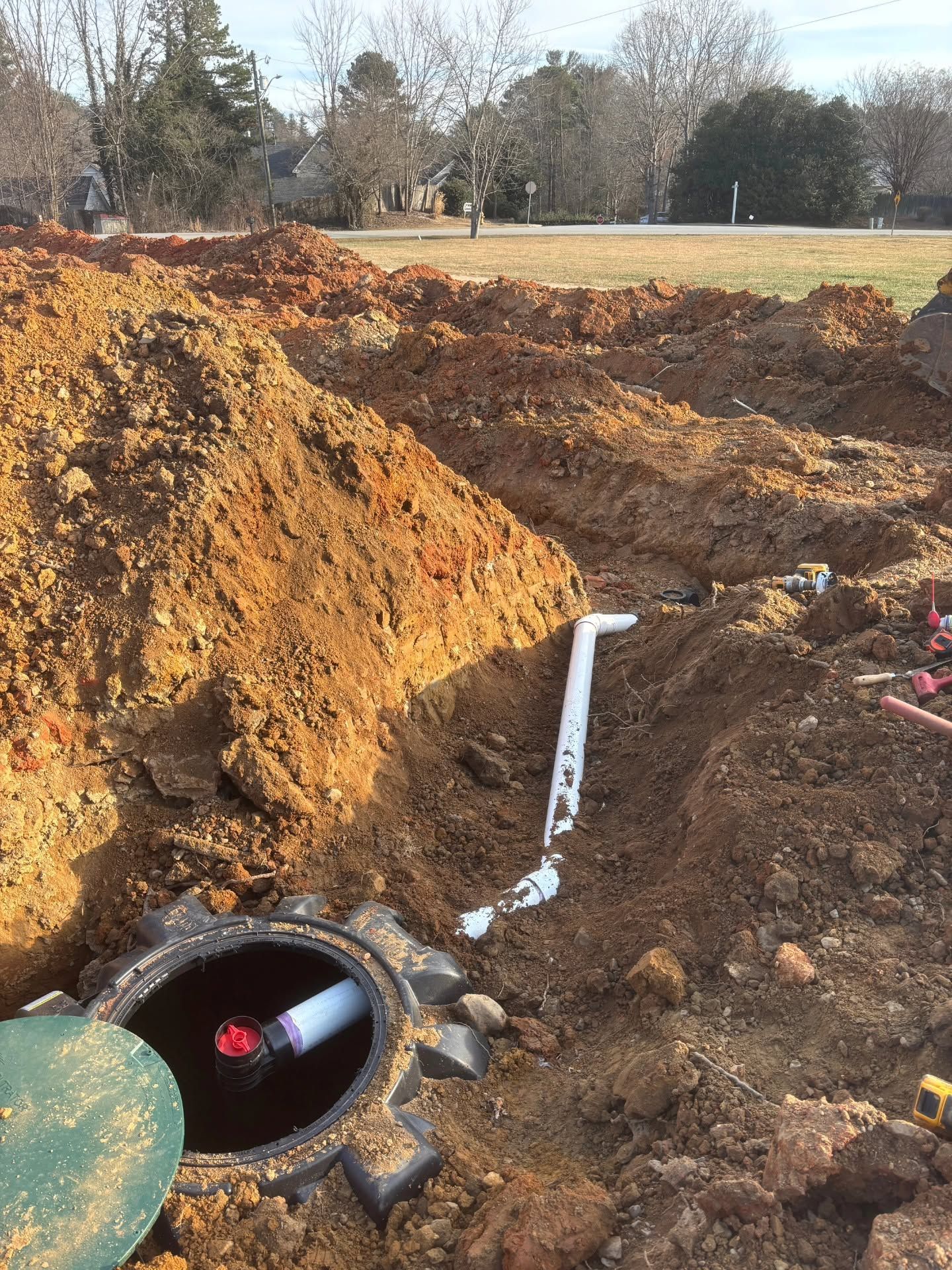 Excavation site with a large hole, exposed orange soil, and a black pipe with a white strap in the foreground