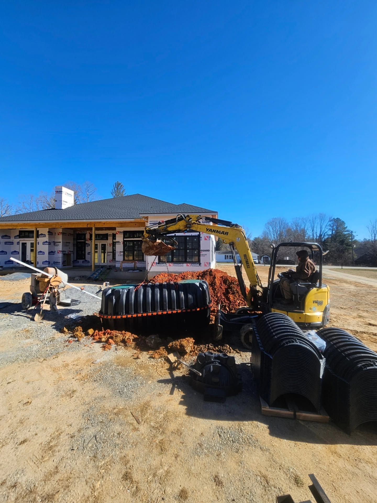 Construction site with excavator trenching near a house, black drainage pipes, and a bright blue sky.