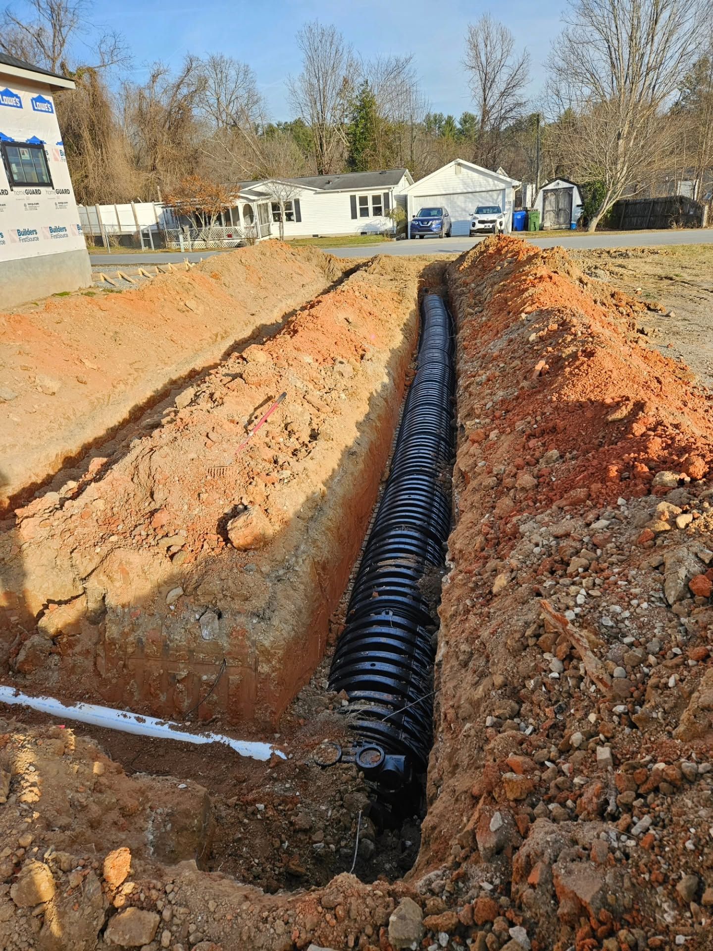 Trench with black drainage pipe laid in dirt at a construction site under clear daylight