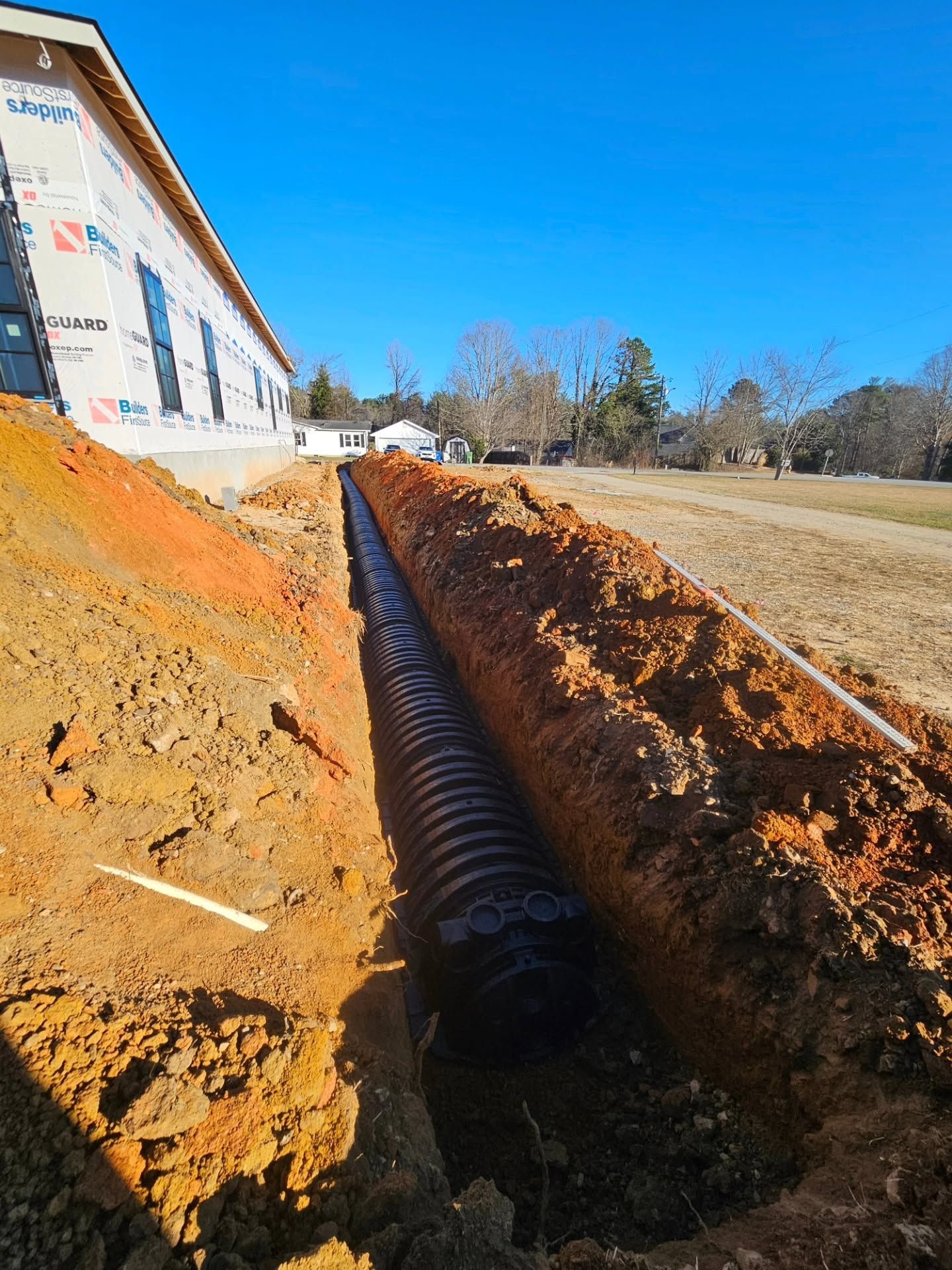 Black drainage pipe in an orange dirt trench beside a building under a clear blue sky