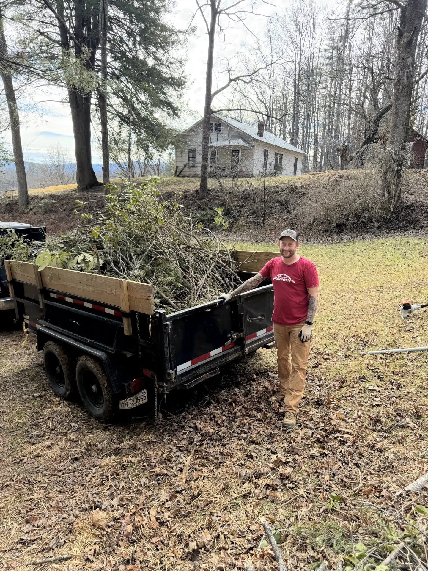 Person standing beside a small trailer full of yard debris in a wooded yard