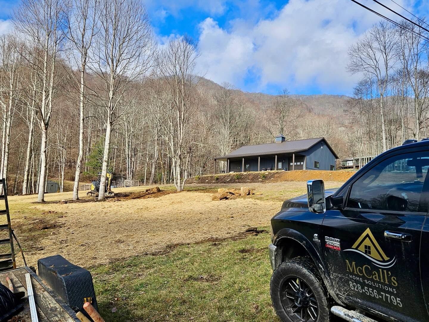 Black pickup by a cabin in a grassy field with bare trees and mountains under a blue sky