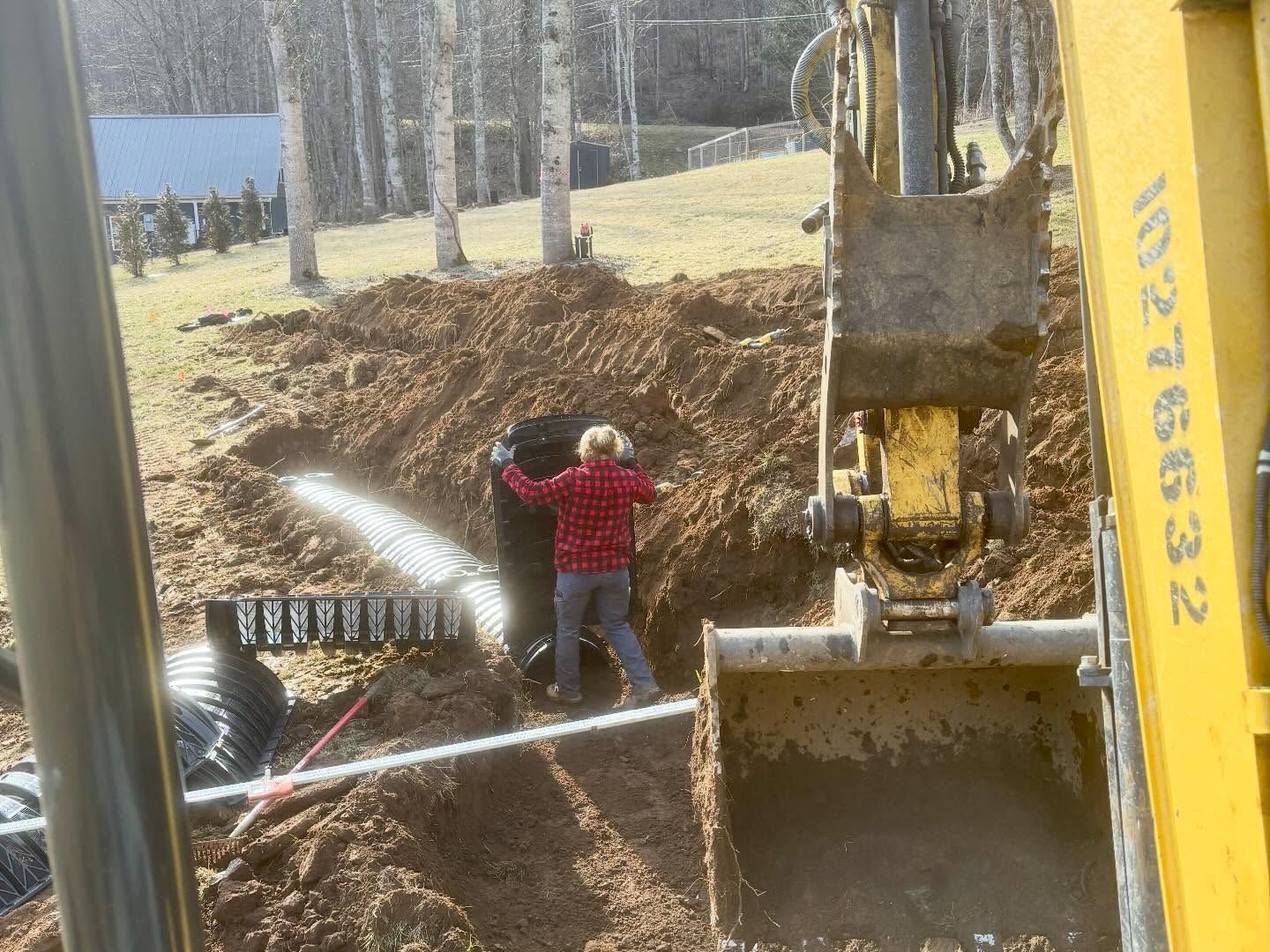 Construction worker standing by excavator, spraying water on a dirt trench near a wooded area