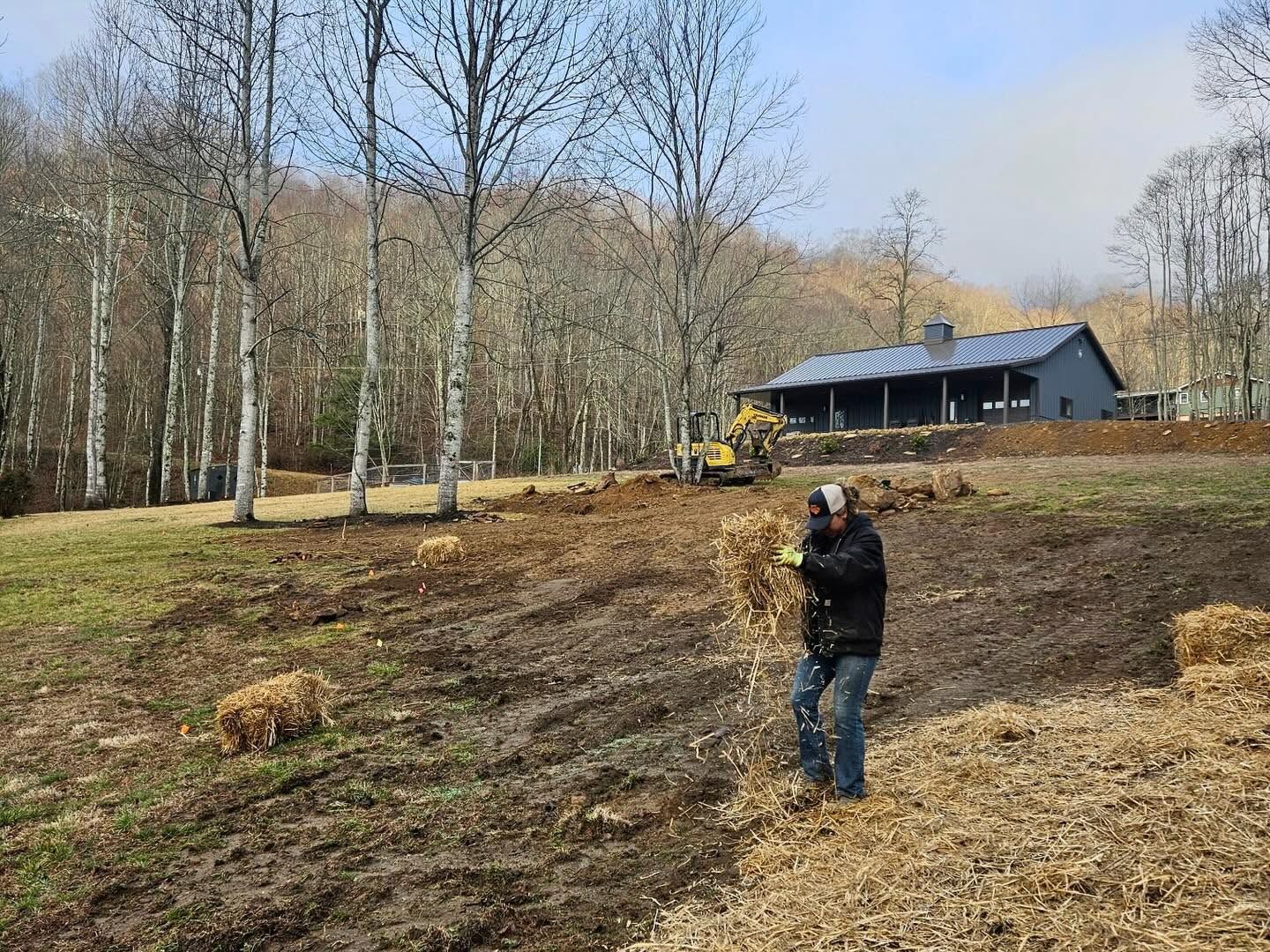 Person standing in a muddy field near a house and a small excavator among leafless trees.