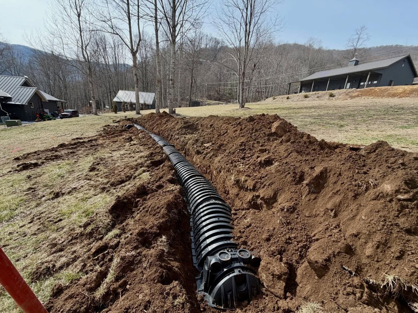 Black drainage pipe laid in a dirt trench across a grassy field near houses and trees