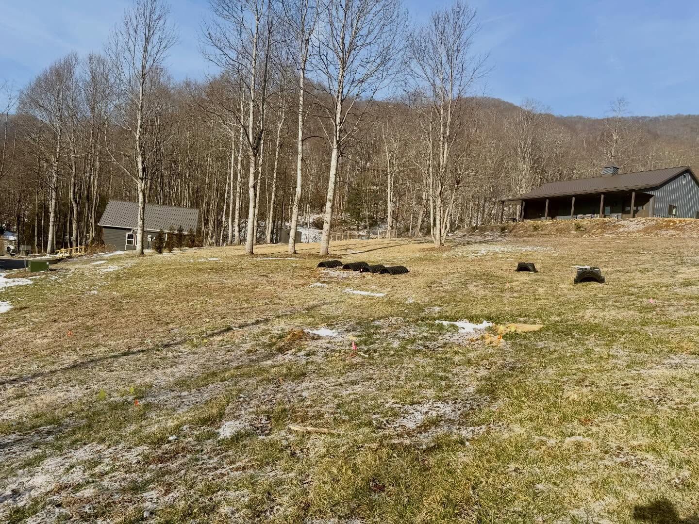 Open grassy field with patches of snow, leafless trees, and two small buildings under a clear sky