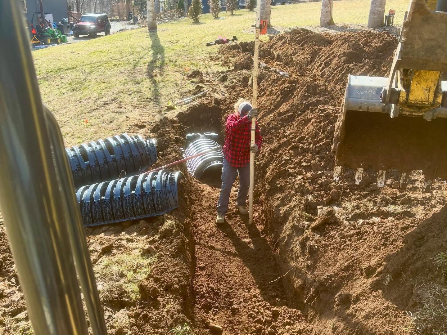 Worker in red jacket stands beside a trench at a construction site with pipes and an excavator.