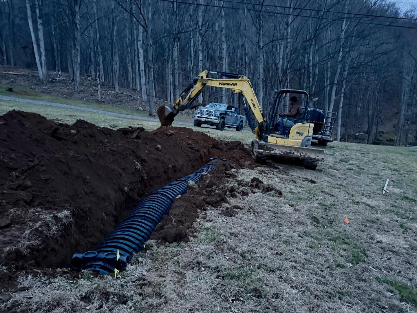 Excavator trenching beside black drainage pipe in a grassy field with wooded background
