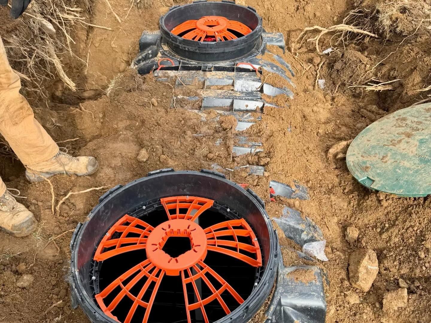 Two orange-and-black pipe access covers installed in a dirt trench, with bricks around them