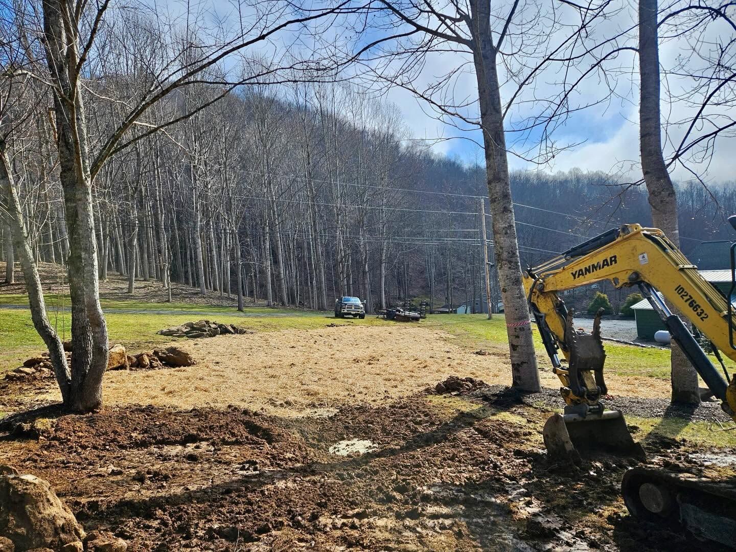 Yellow excavator clearing a grassy hillside with leafless trees and mountains in the background