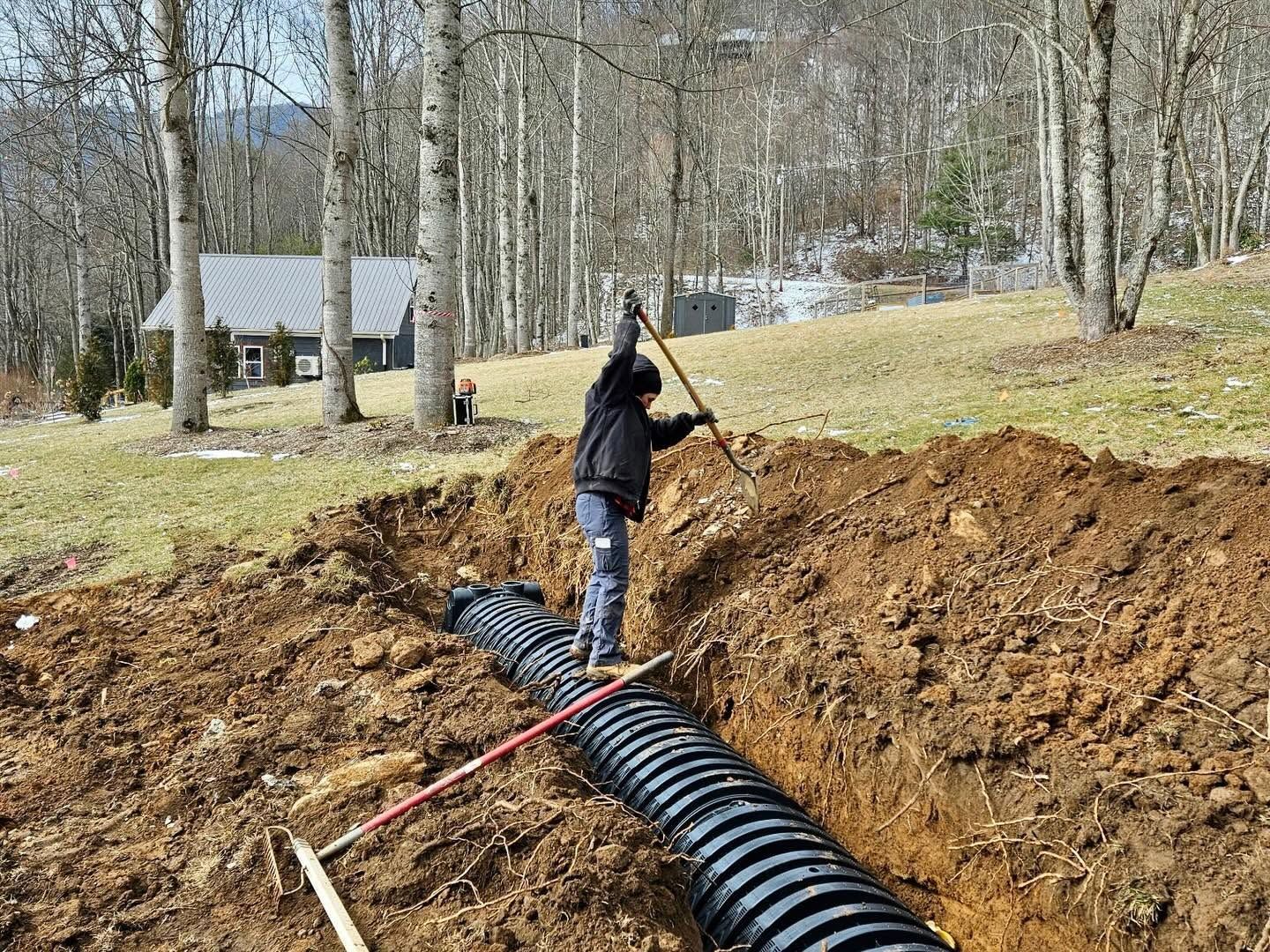 Person shoveling dirt beside a large black drainage pipe in a wooded yard