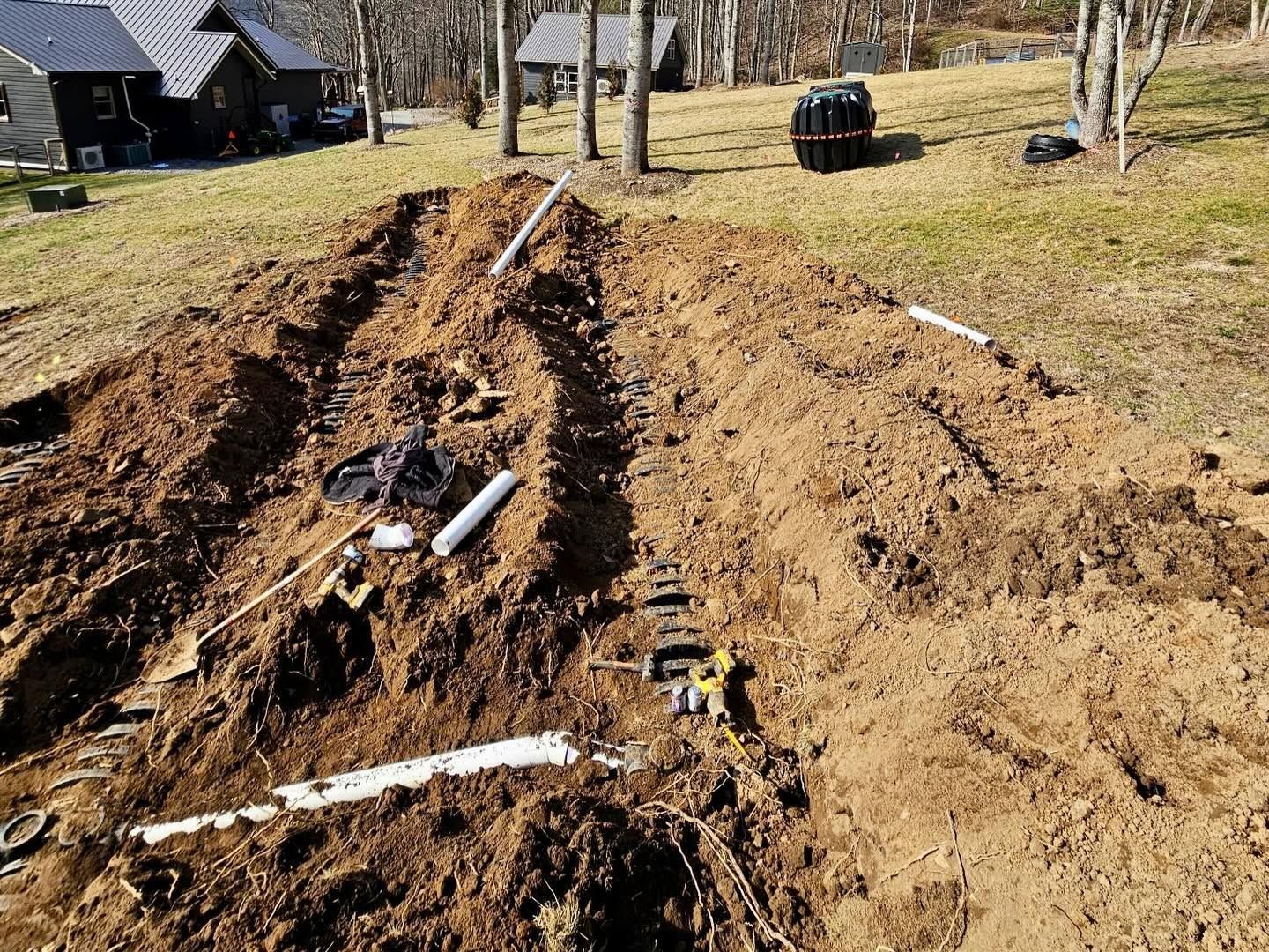 Trenched dirt yard with white pipes and tools laid out beside a house in a rural area