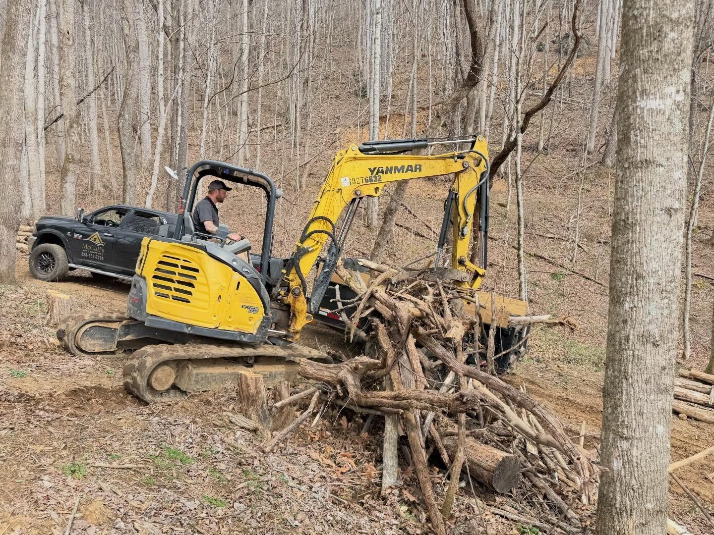 Yellow excavator clearing a pile of tree branches on a wooded hillside next to a black SUV