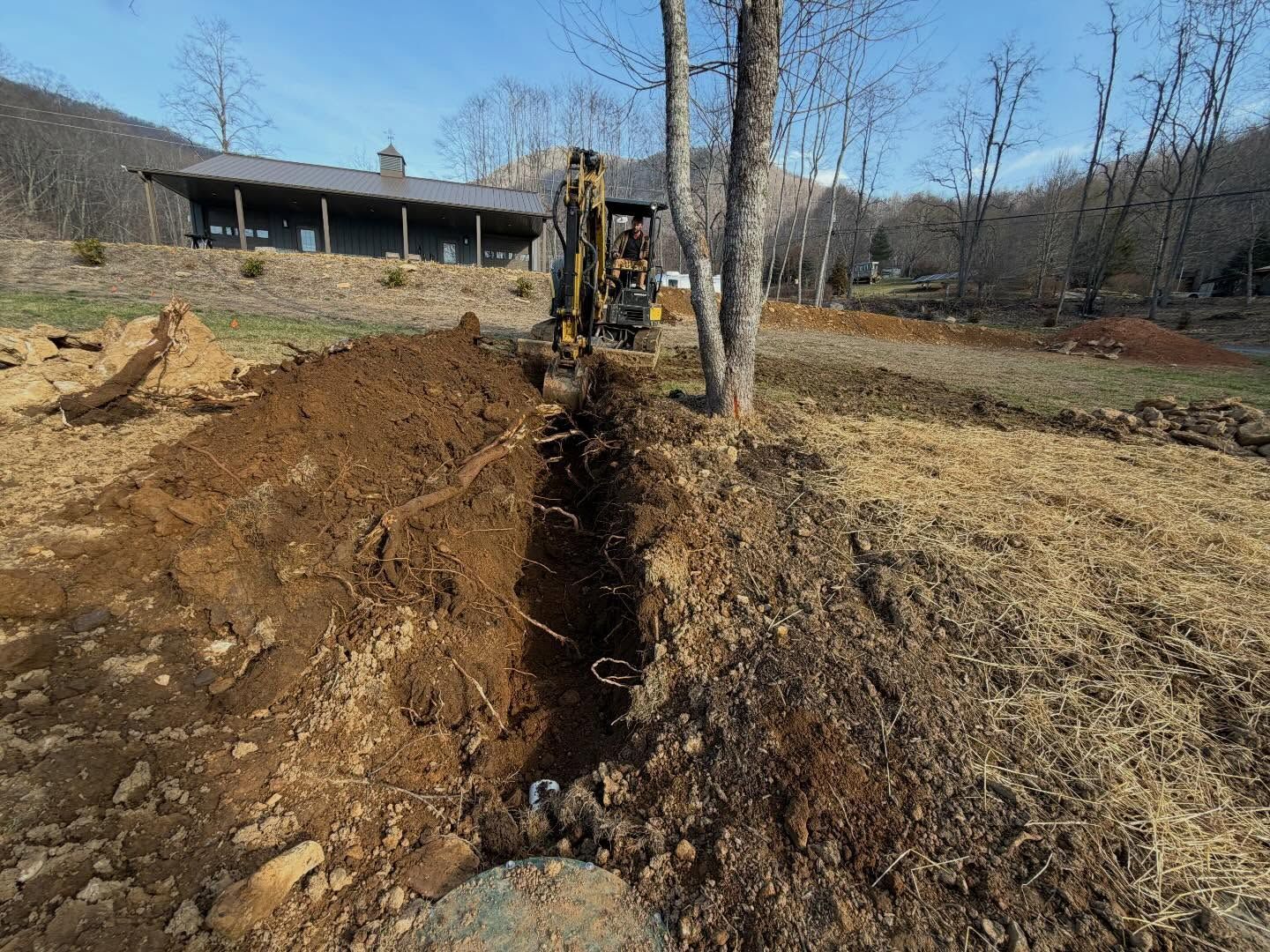 Excavator digging a trench through a dirt yard near a house and bare trees