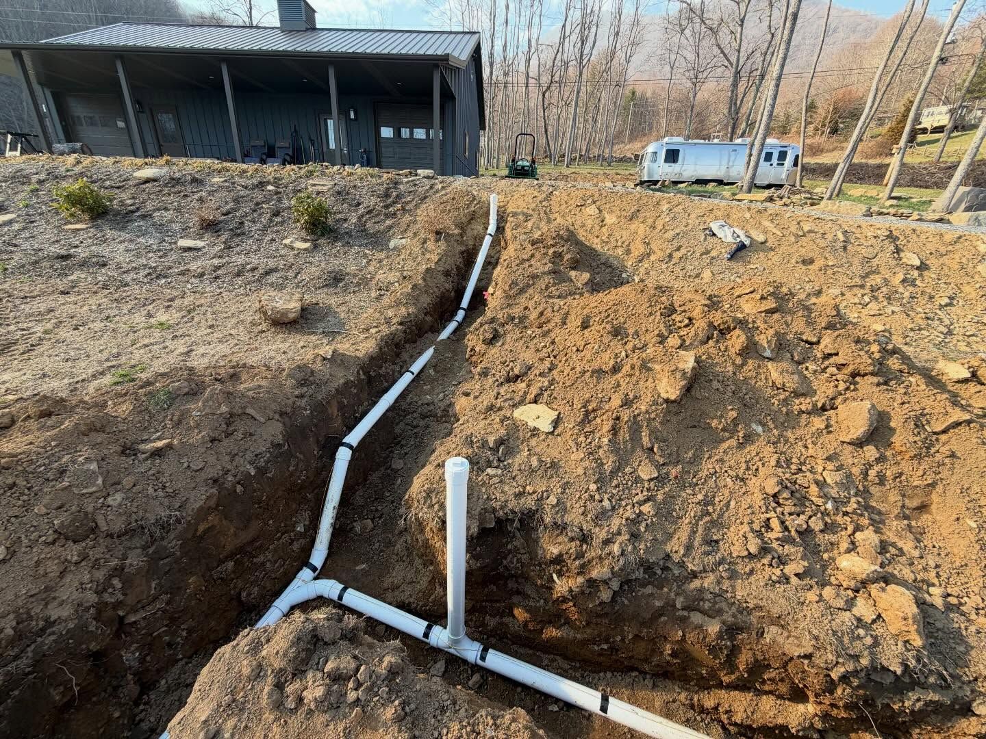 Trench with white pipes running through a dirt yard near a house and trees