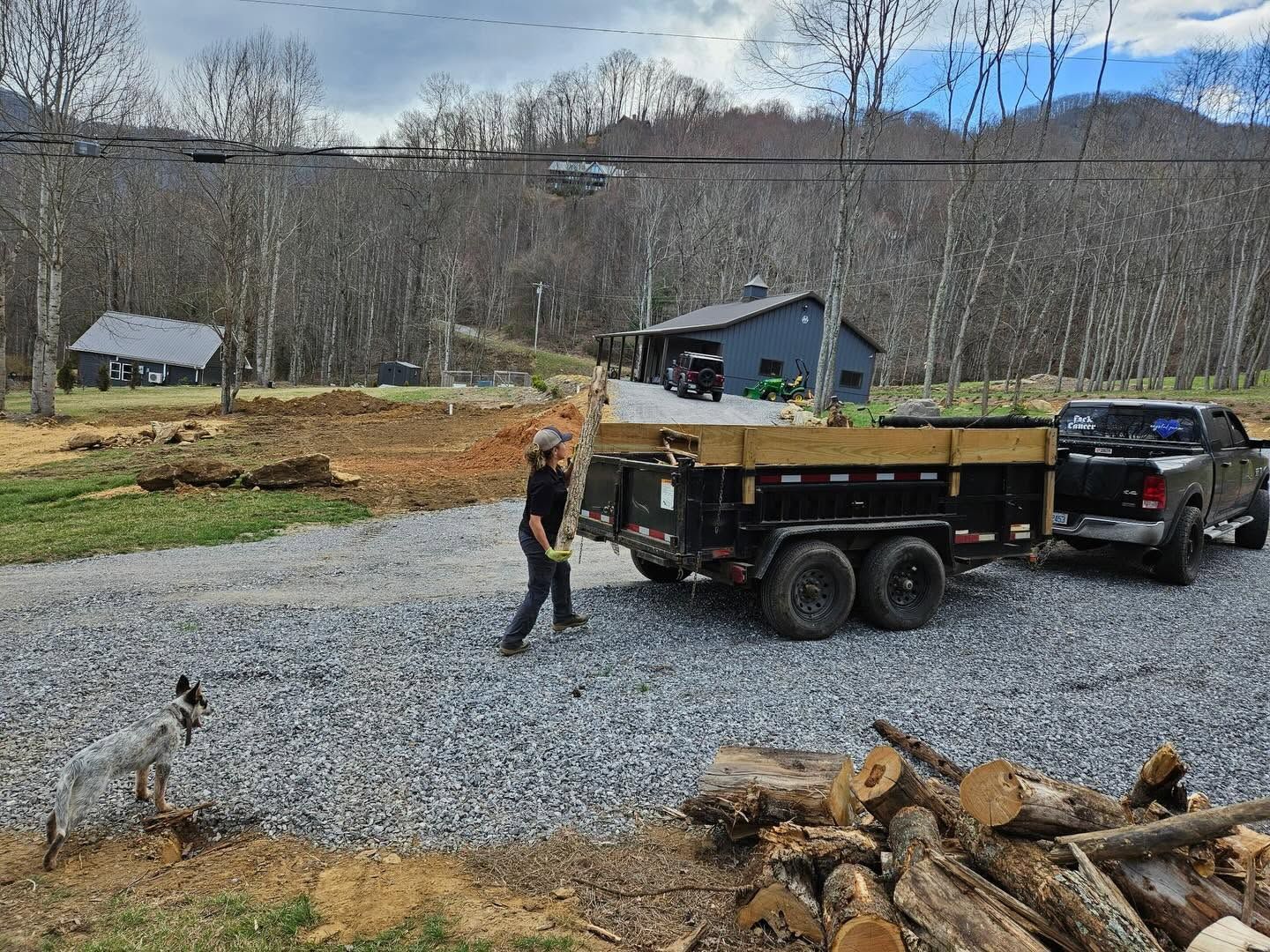 Person beside a gravel-filled trailer hitched to a truck in a rural yard, with cut logs in the foreground.