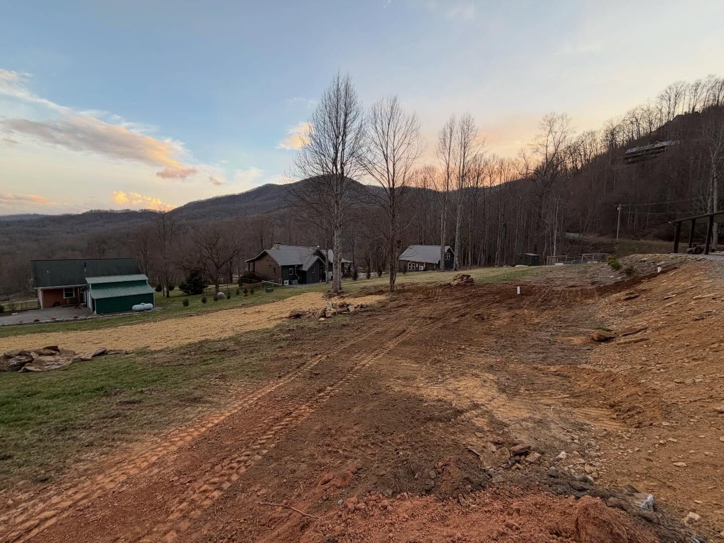 Rural dirt path at sunset with bare trees, small buildings, and distant hills under a pastel sky