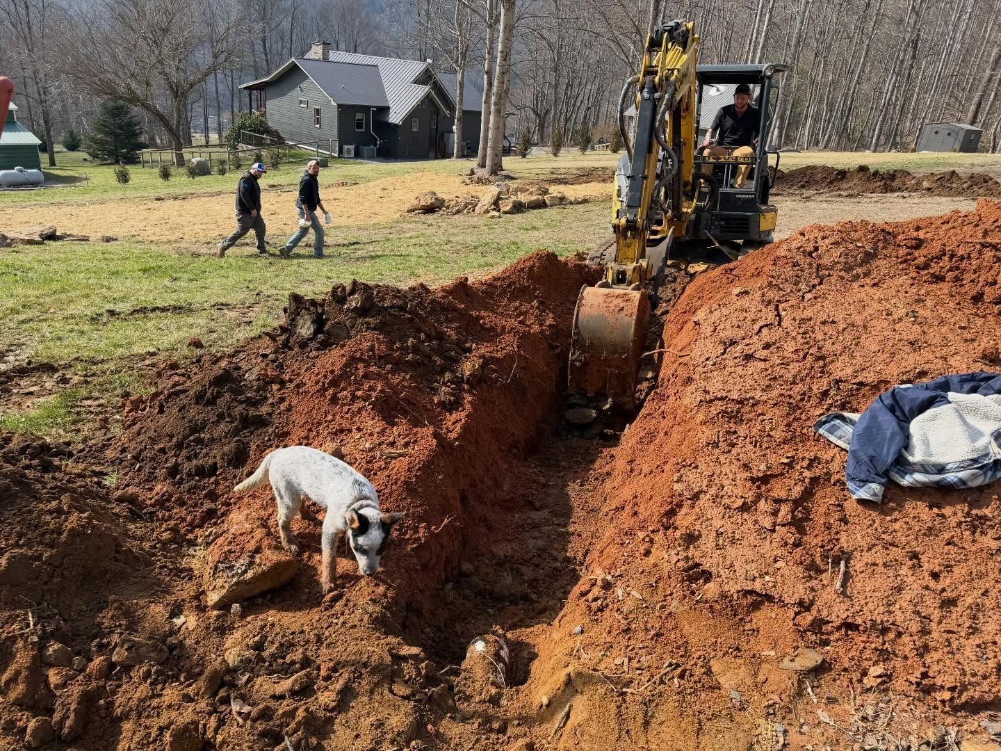 Workers and a backhoe digging a large trench in a red dirt field near a house