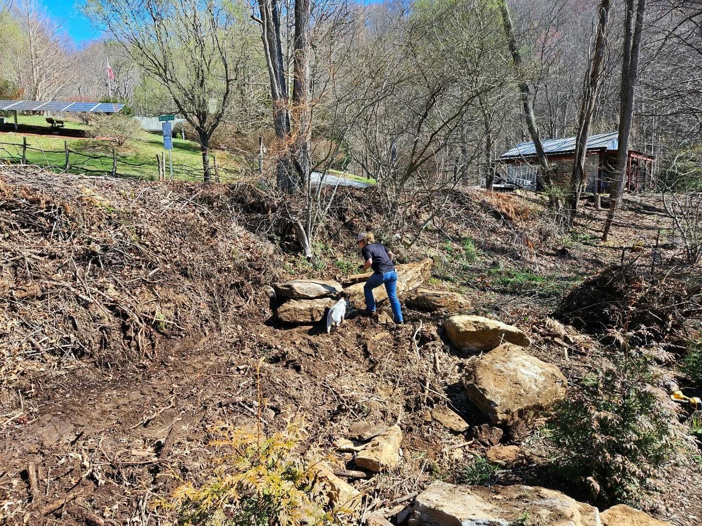 Person in blue working on a rocky creek bank in a wooded area under a clear sky