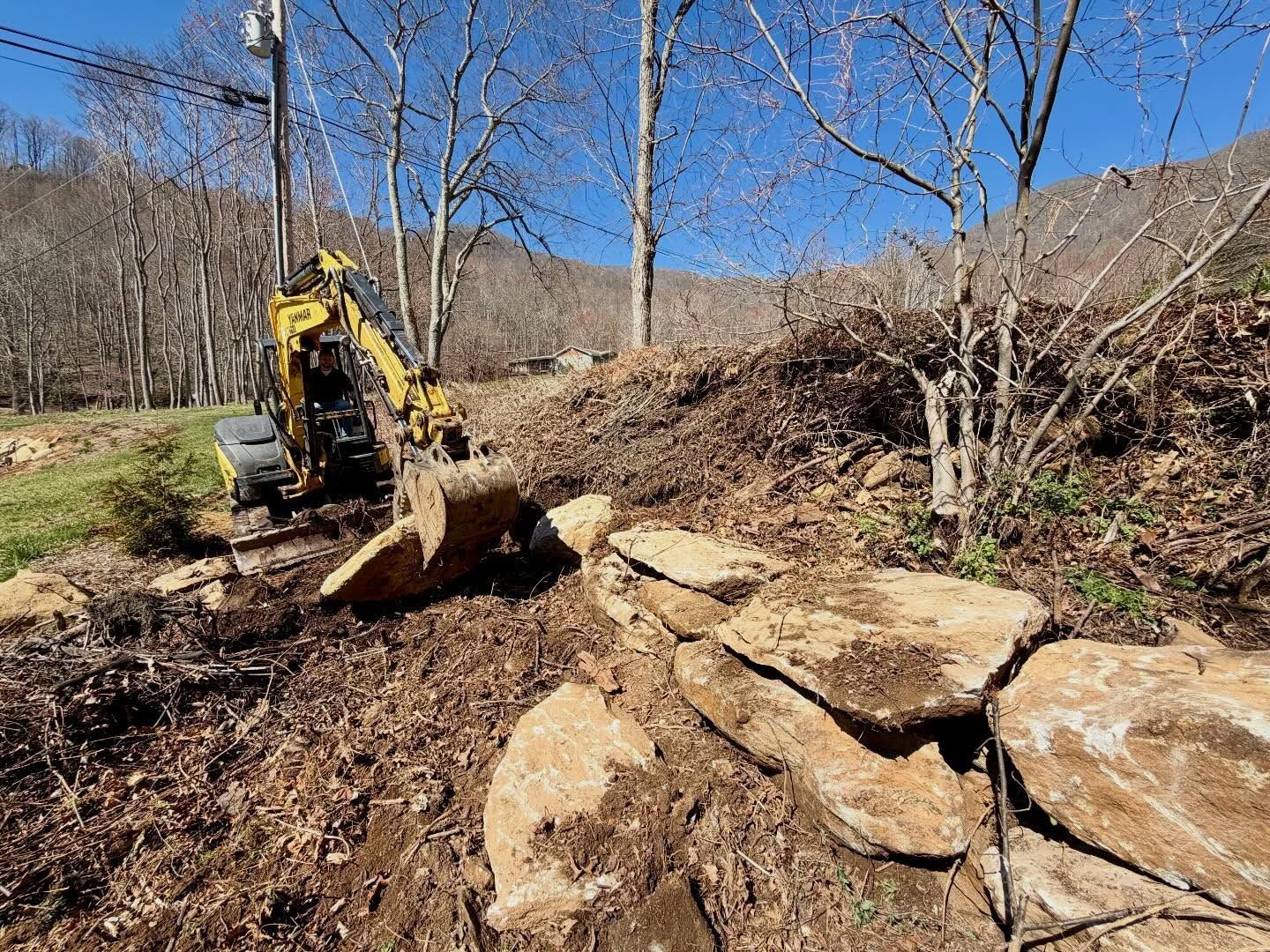 Yellow excavator clearing a rocky, leaf-covered hillside under a blue sky