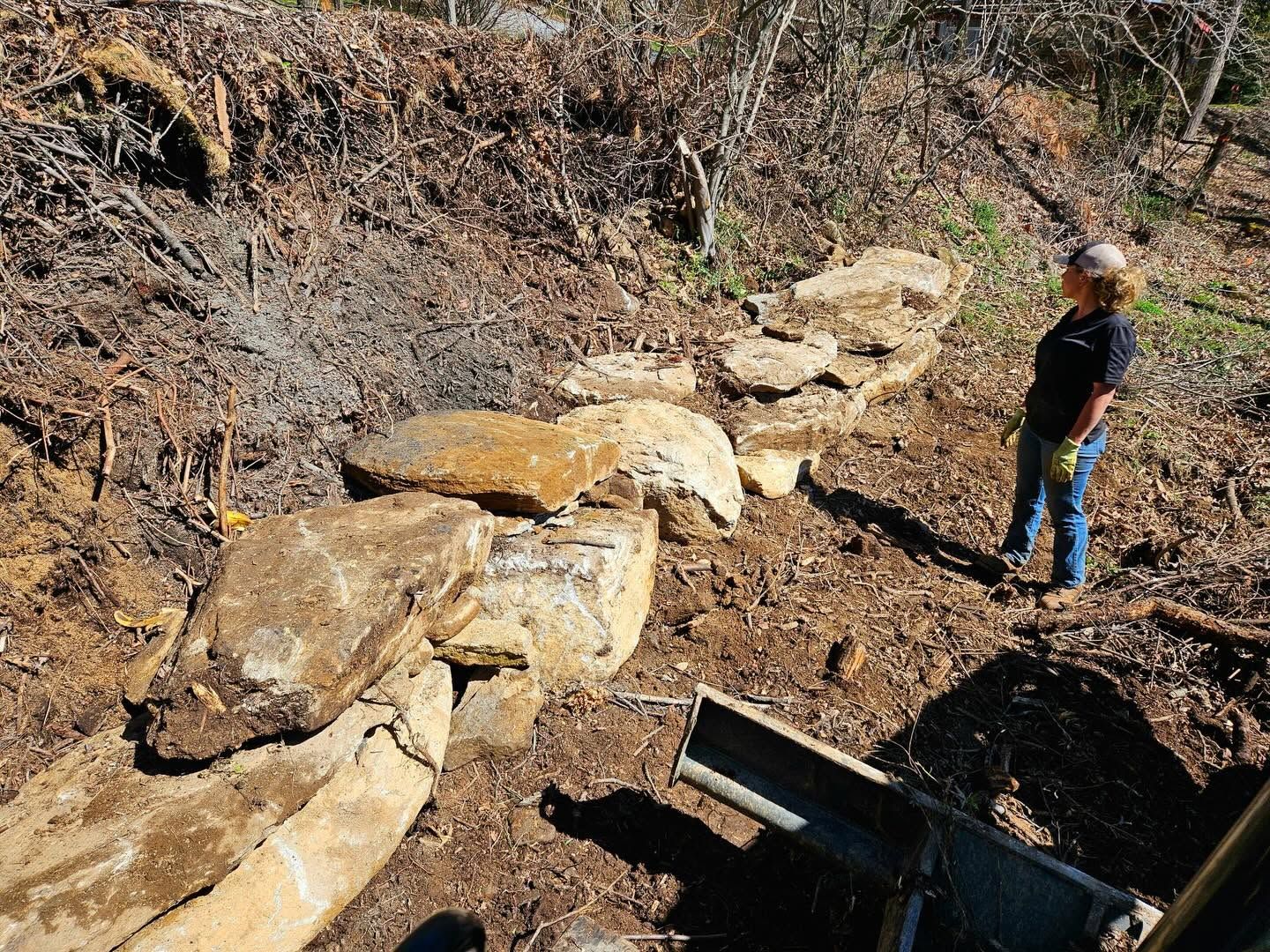Person standing near large rocks on a wooded hillside beside a small bridge or culvert.