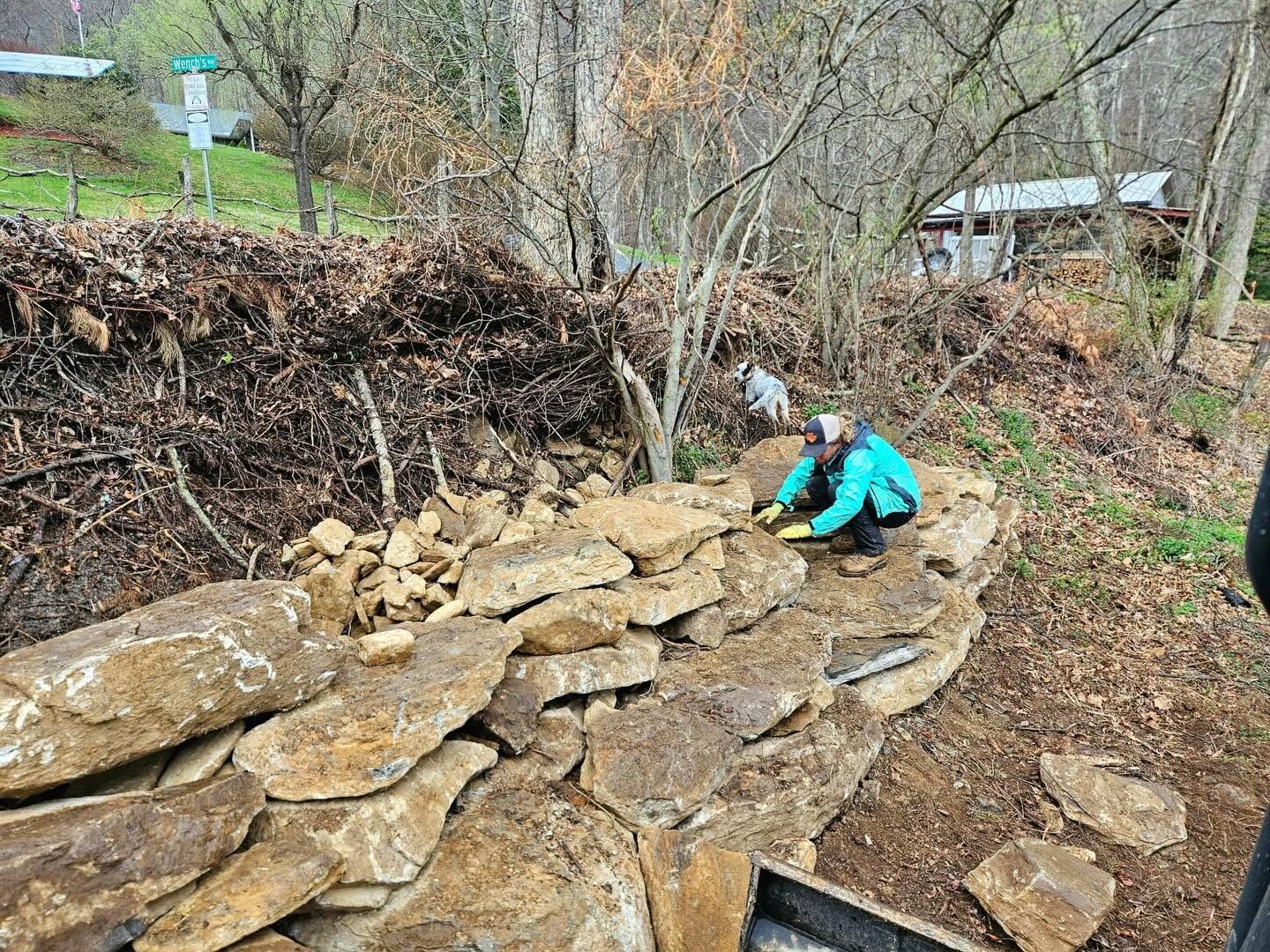 Person in teal jacket examining a stone retaining wall on a wooded hillside.