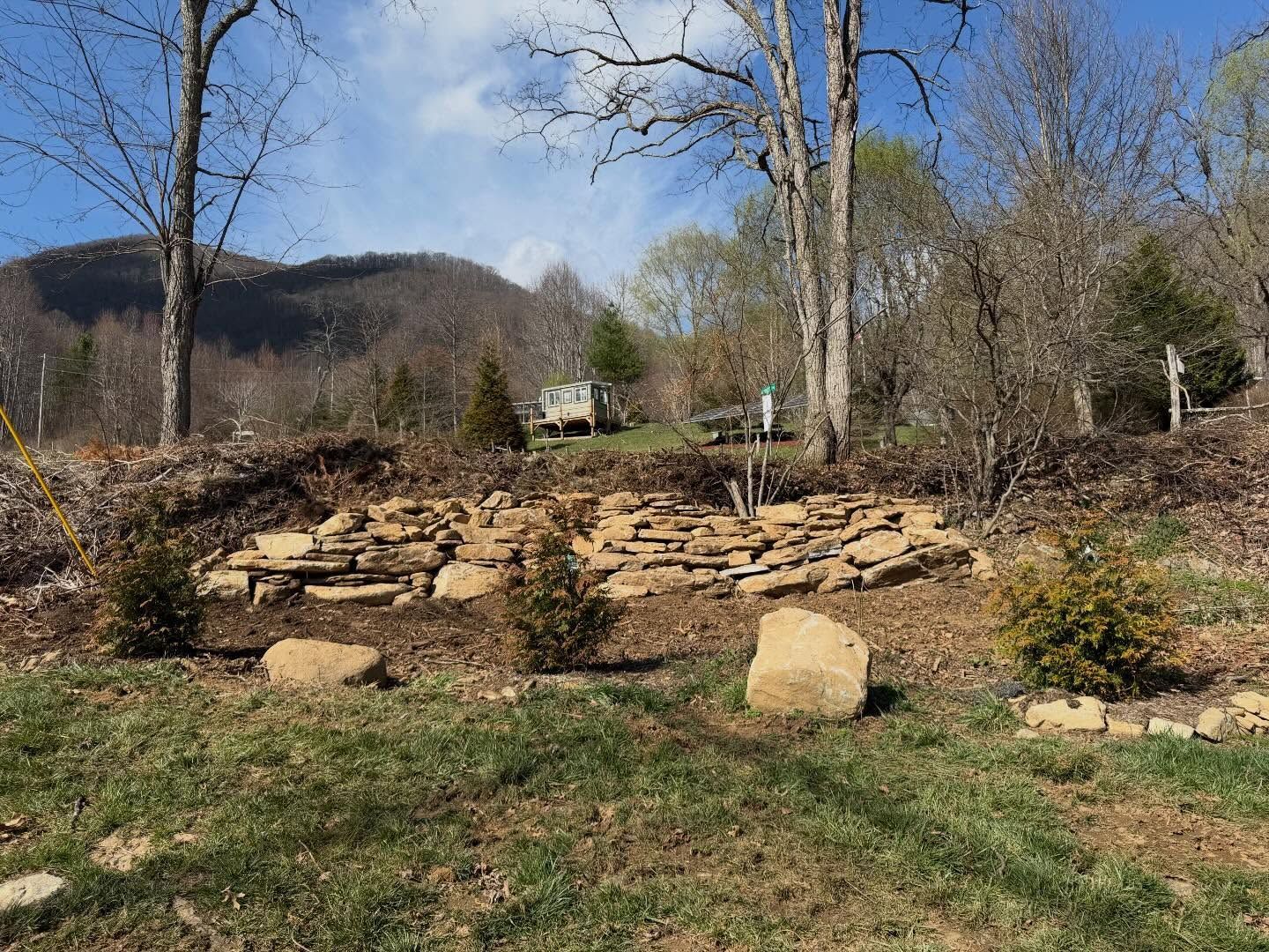 Stone garden wall in a wooded yard with a grassy foreground and hills under a blue sky