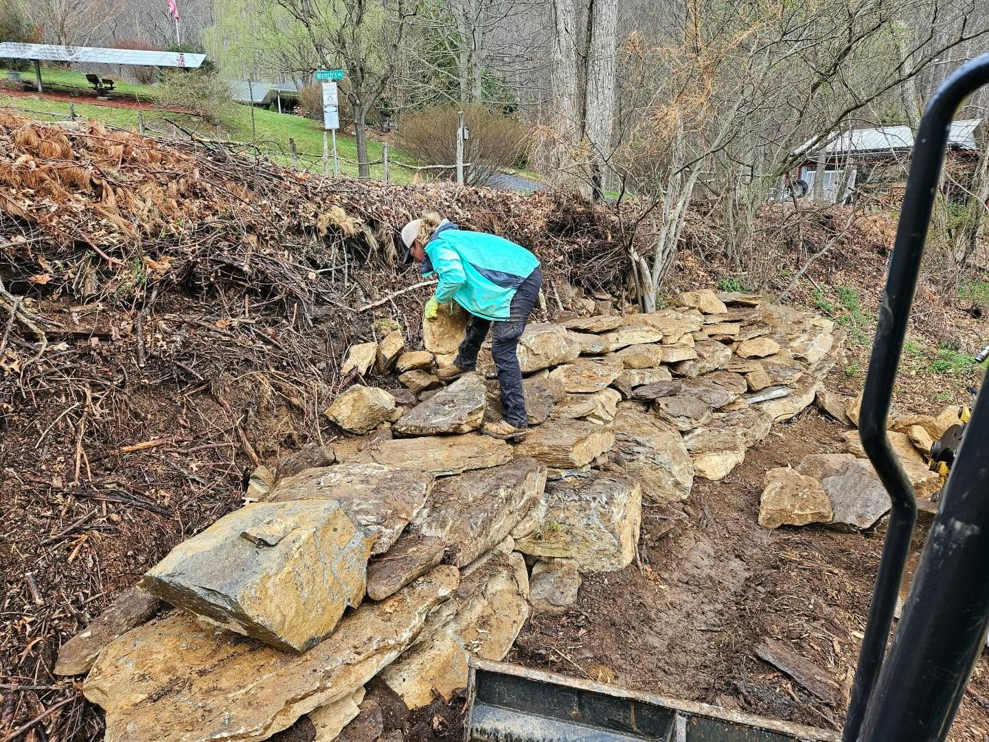 Person in a turquoise jacket stacking rocks on a stone wall in a wooded yard