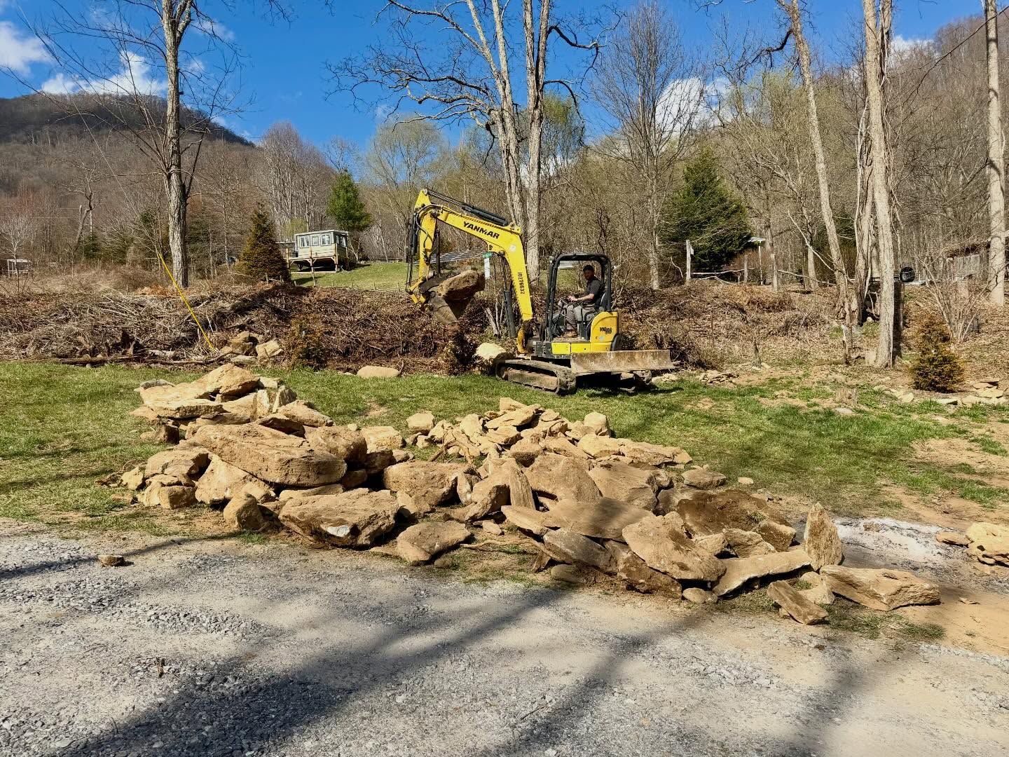 Yellow excavator clearing brush beside a stone pile in a wooded rural area under a blue sky