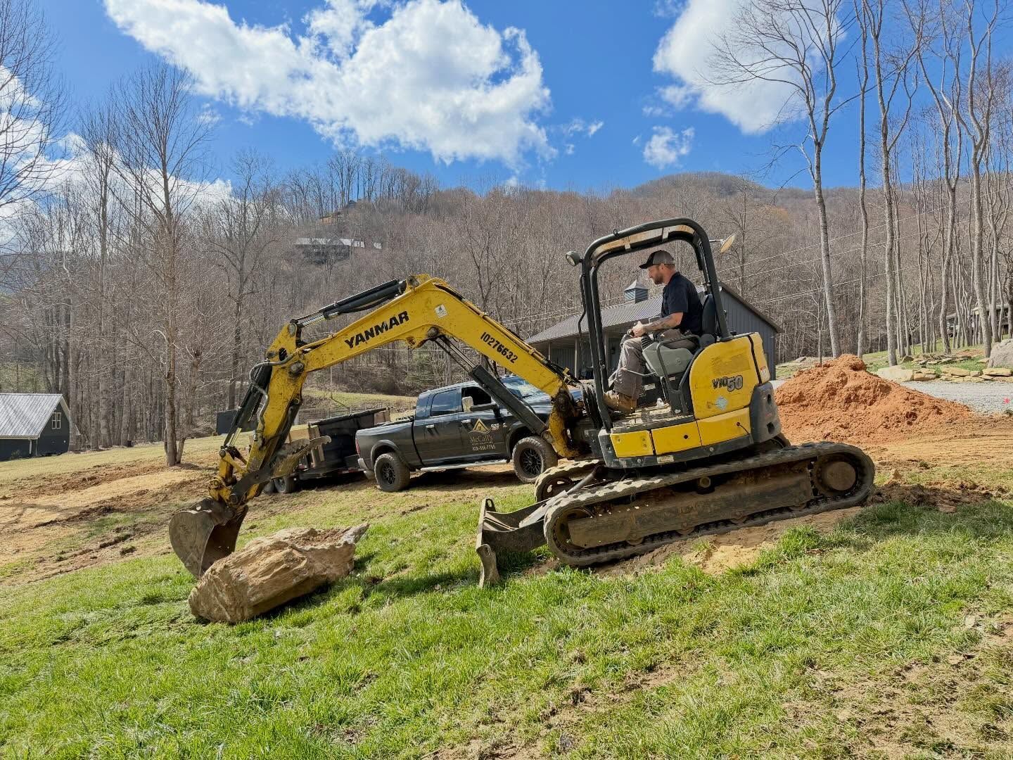 Yellow excavator moving a large rock in a grassy yard with a pickup truck and hills in the background