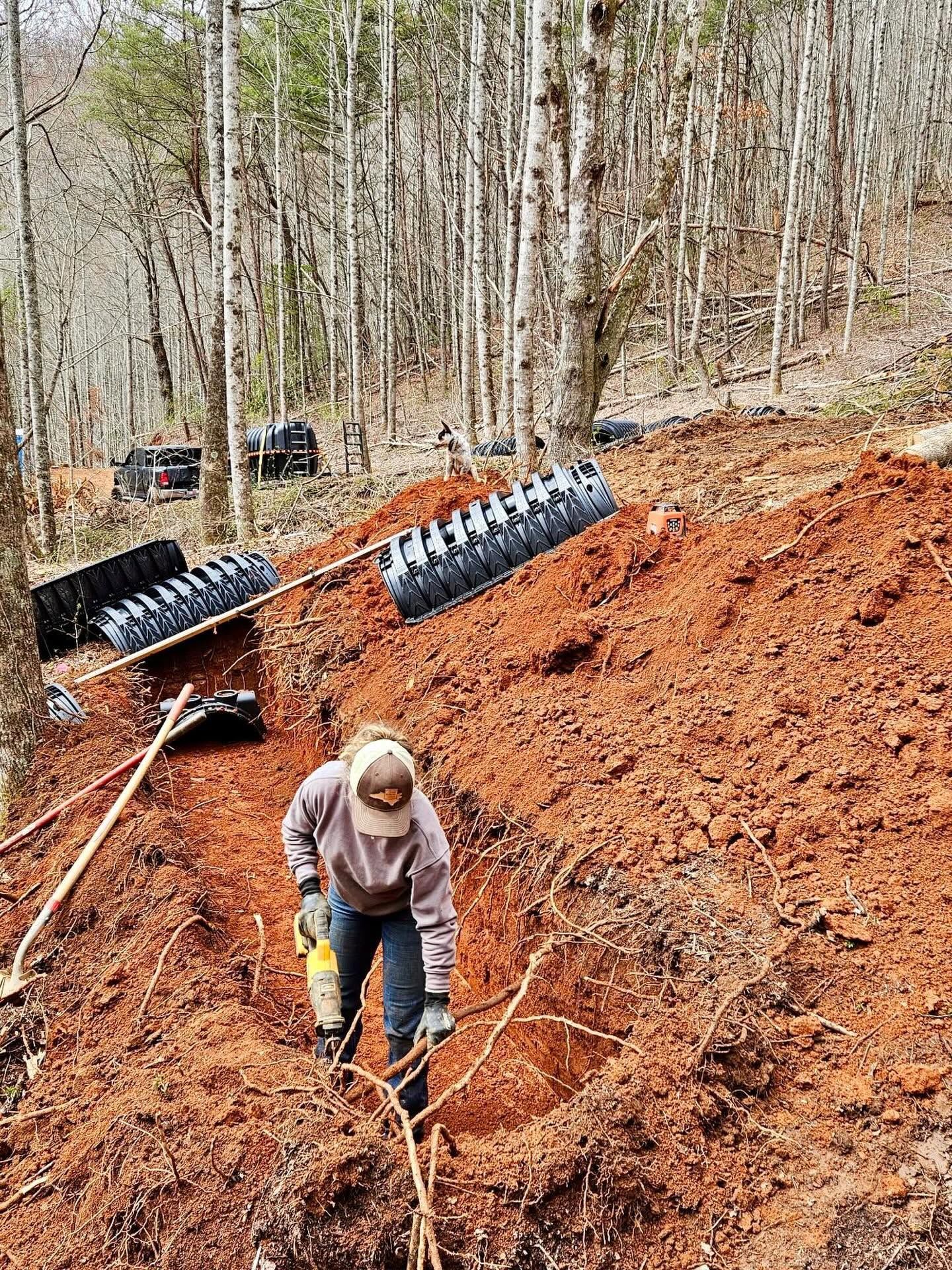 Worker digging on a steep red dirt hillside with erosion control mats and a forest in the background