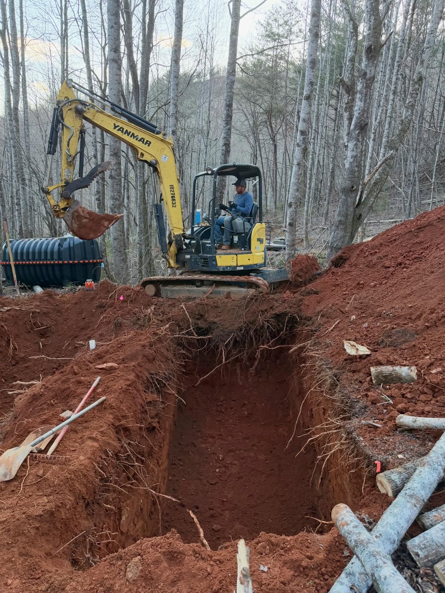 Yellow excavator digging a trench beside a red soil mound in a wooded area