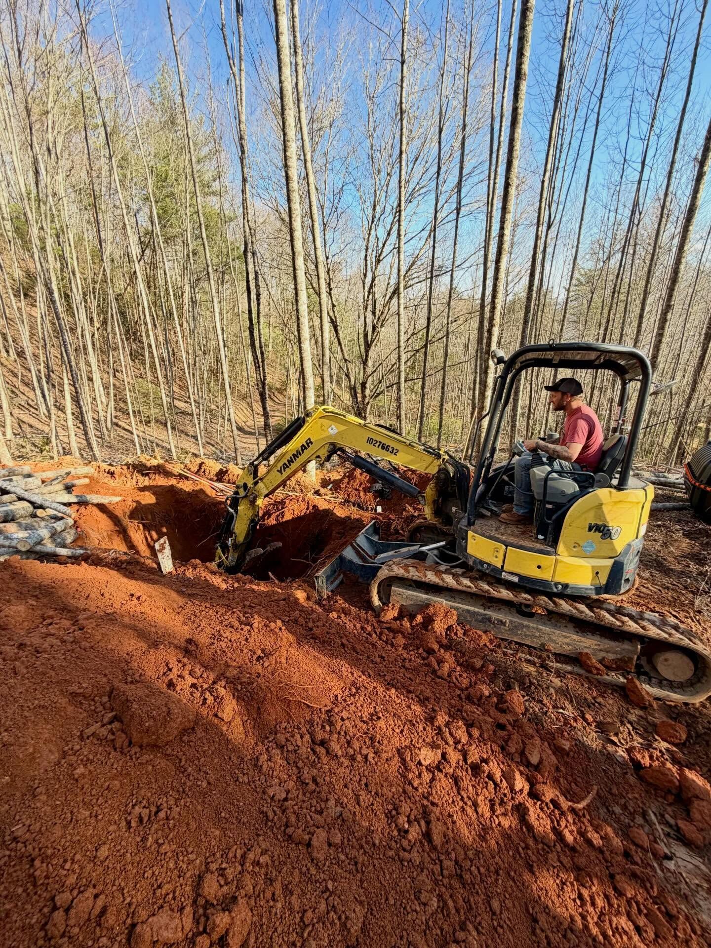 Yellow excavator digging a red dirt trench in a wooded area, with an operator in the cab
