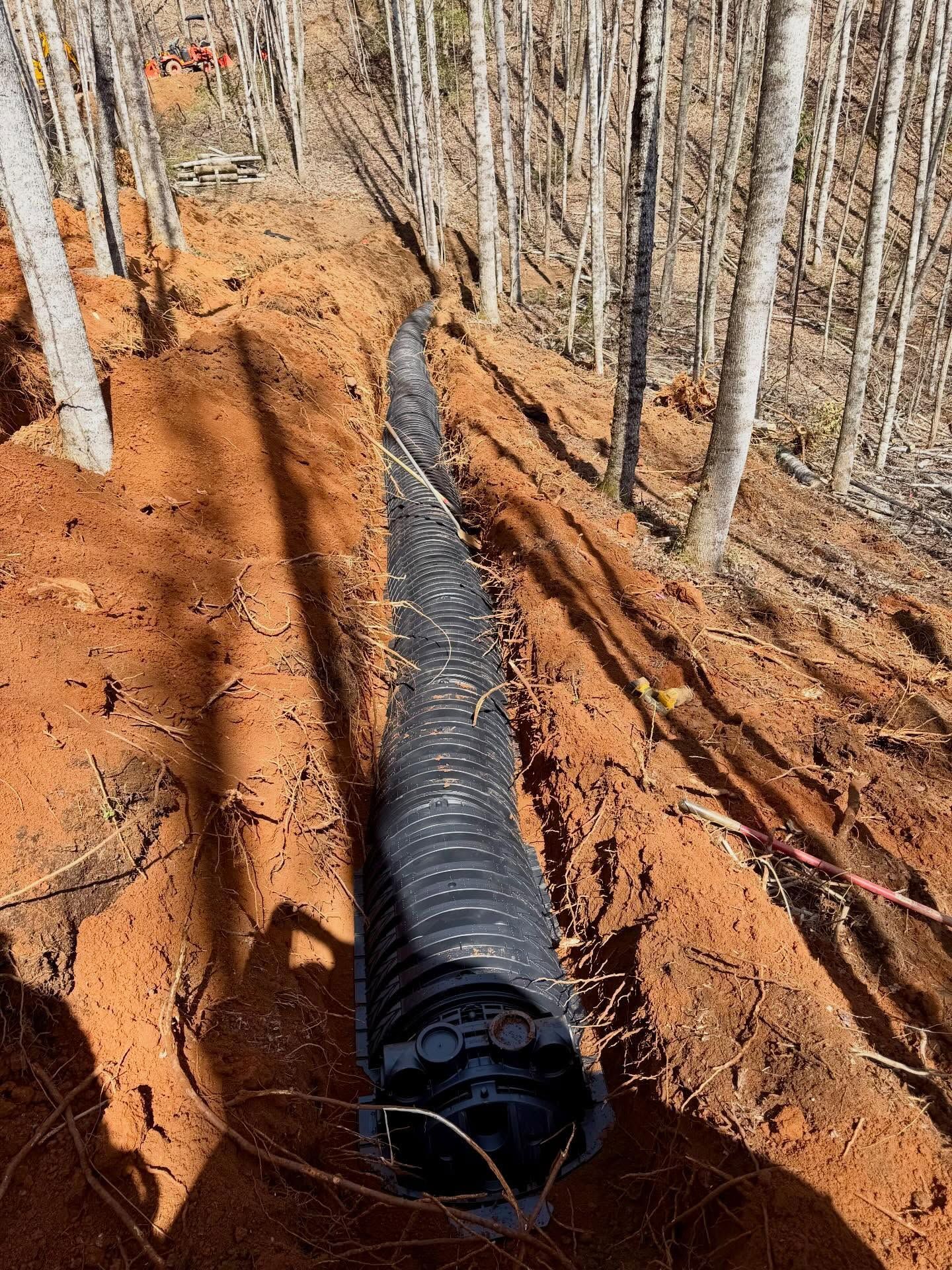 Black drainage pipe laid in a red dirt trench amid a wooded hillside with bare trees