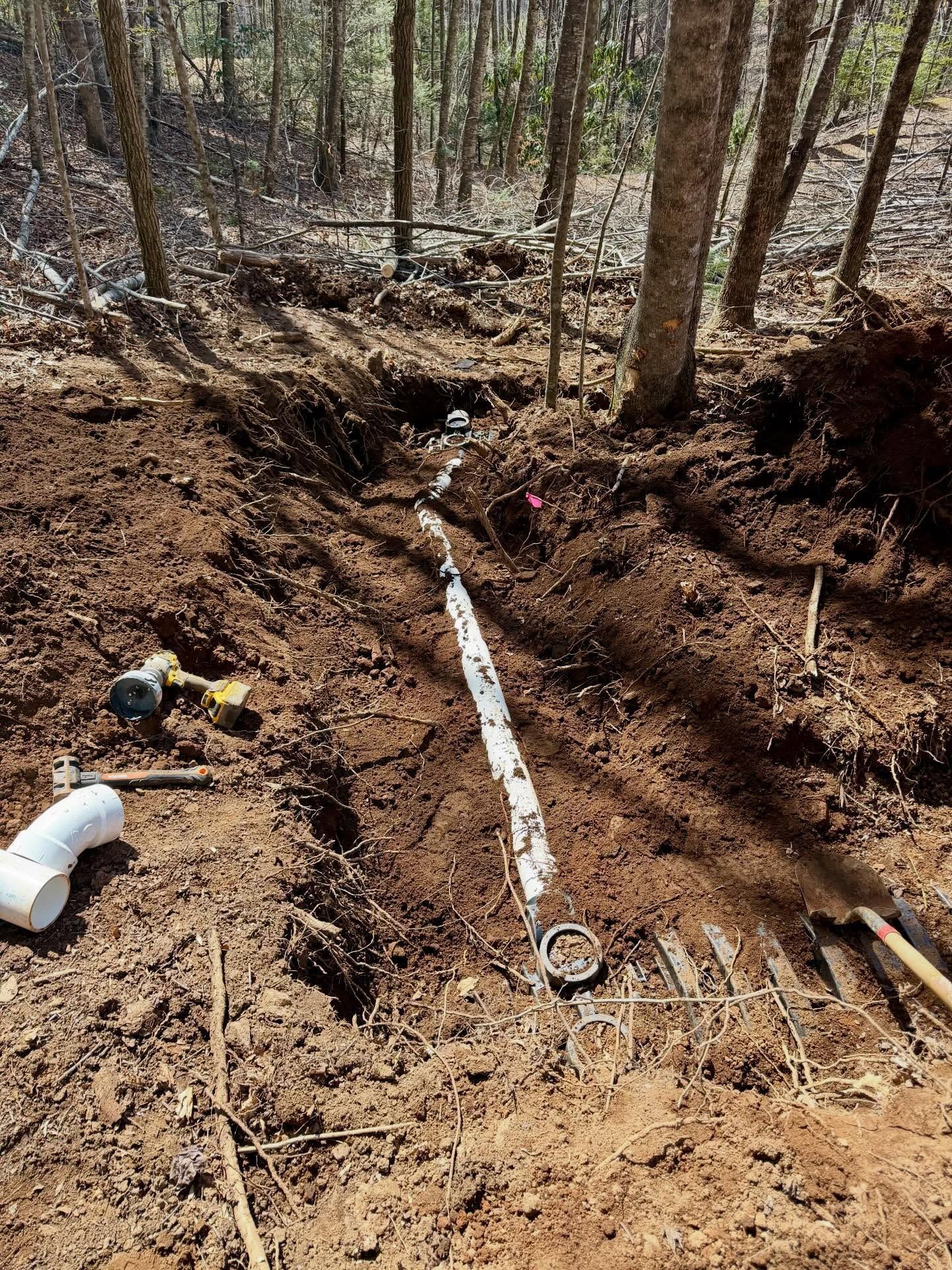 Excavated trench on a wooded hillside with exposed white pipe and dirt work area