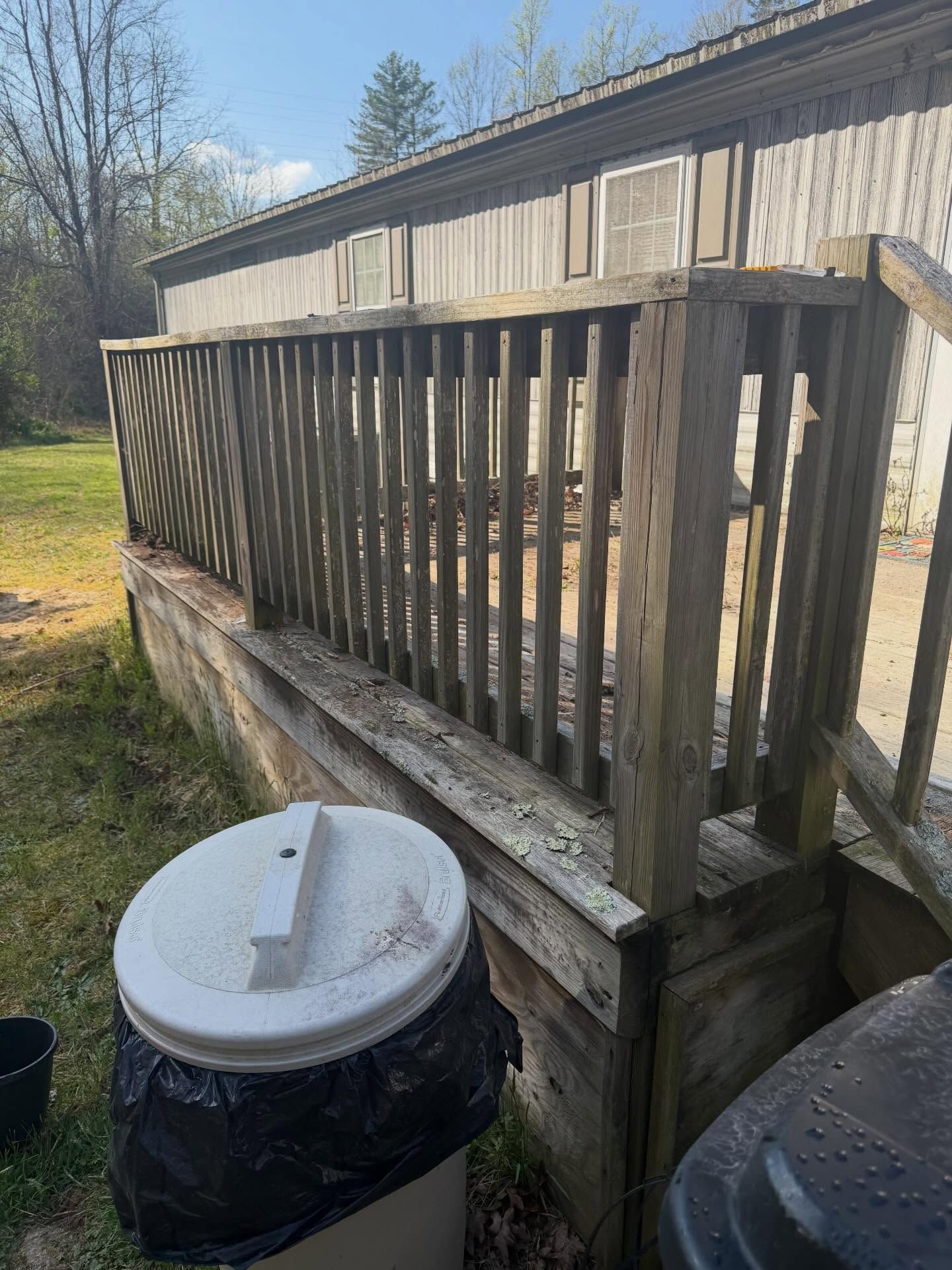 Weathered wooden porch with railings beside a house, with a trash bin in the foreground and trees around it
