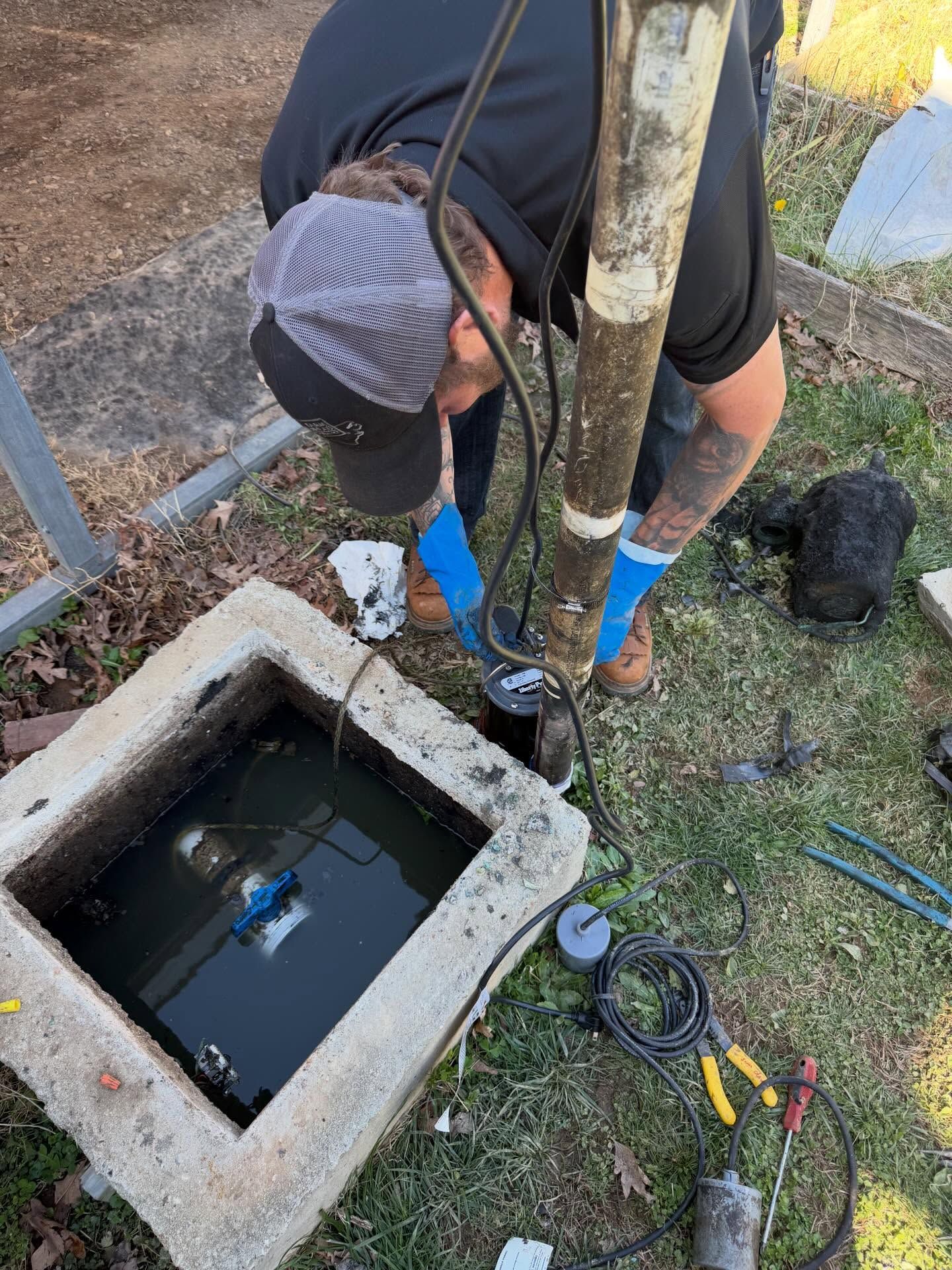 Worker leaning over an open utility box, inspecting wiring next to a pole and muddy ground.