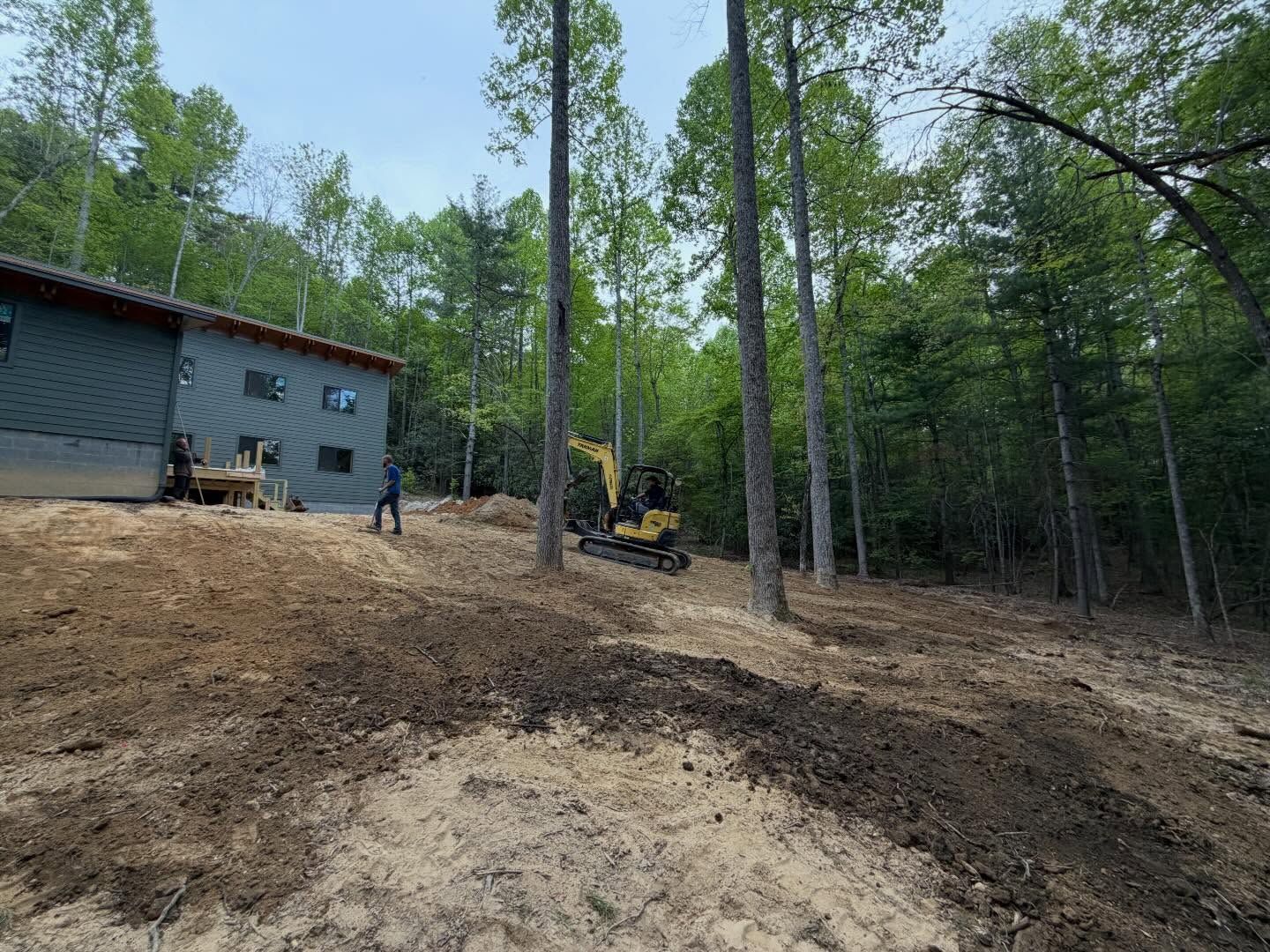 Excavator grading a dirt slope beside a house in a wooded yard.