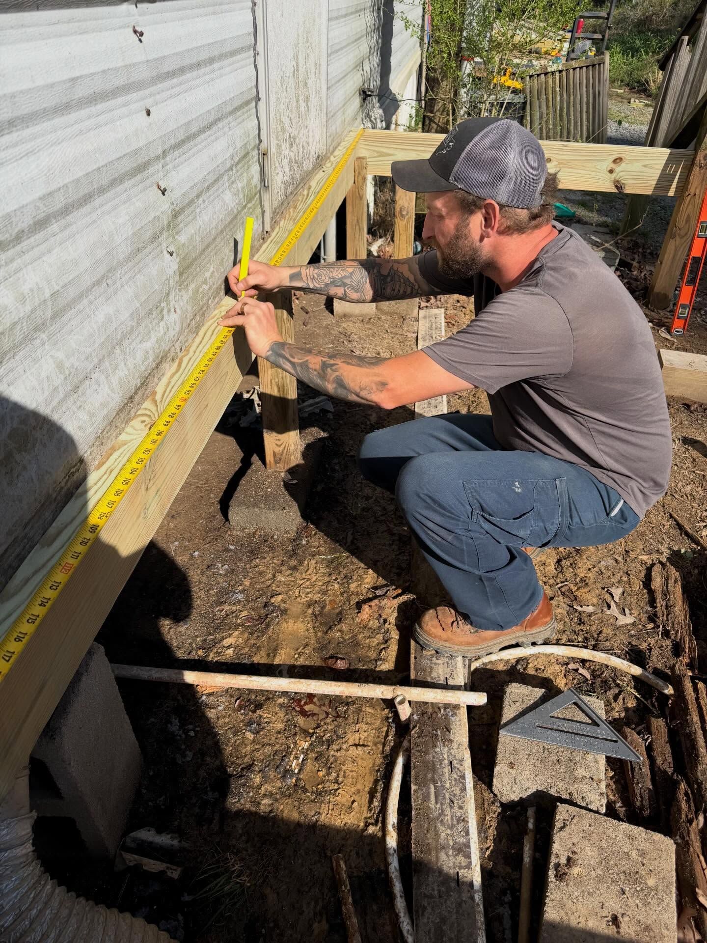 Worker kneeling beside a trailer, measuring along the lower edge with a yellow tape measure outdoors.